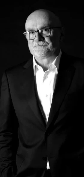 Black and white portrait of a bald man with glasses, wearing a suit and white shirt.