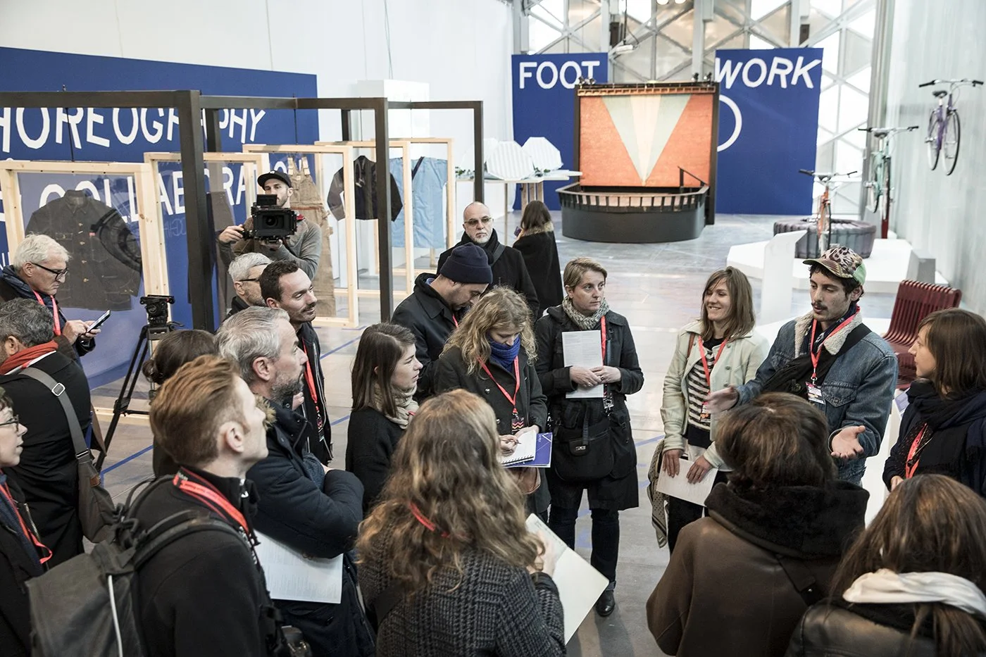 Group of people attending a presentation or discussion at an indoor event, with blue signs reading "FOOT" and "WORK" in the background, and some display items including bicycles and apparel.