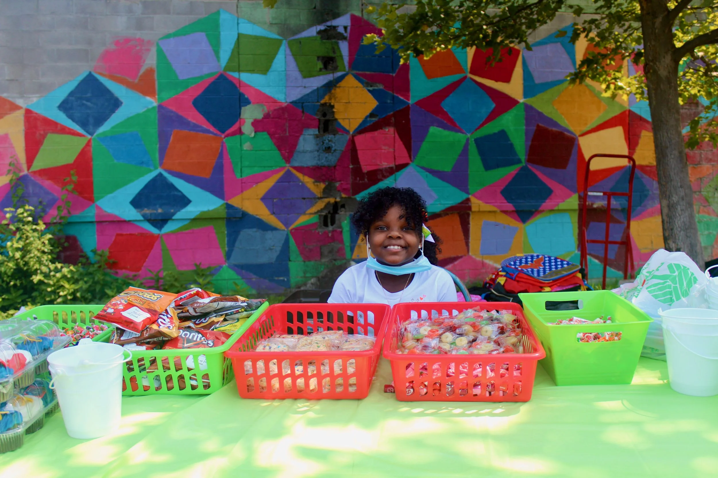 A young girl with curly hair smiles at a table outdoors, selling various snacks sorted in colorful baskets, with a bright, geometric painted mural and trees in the background.