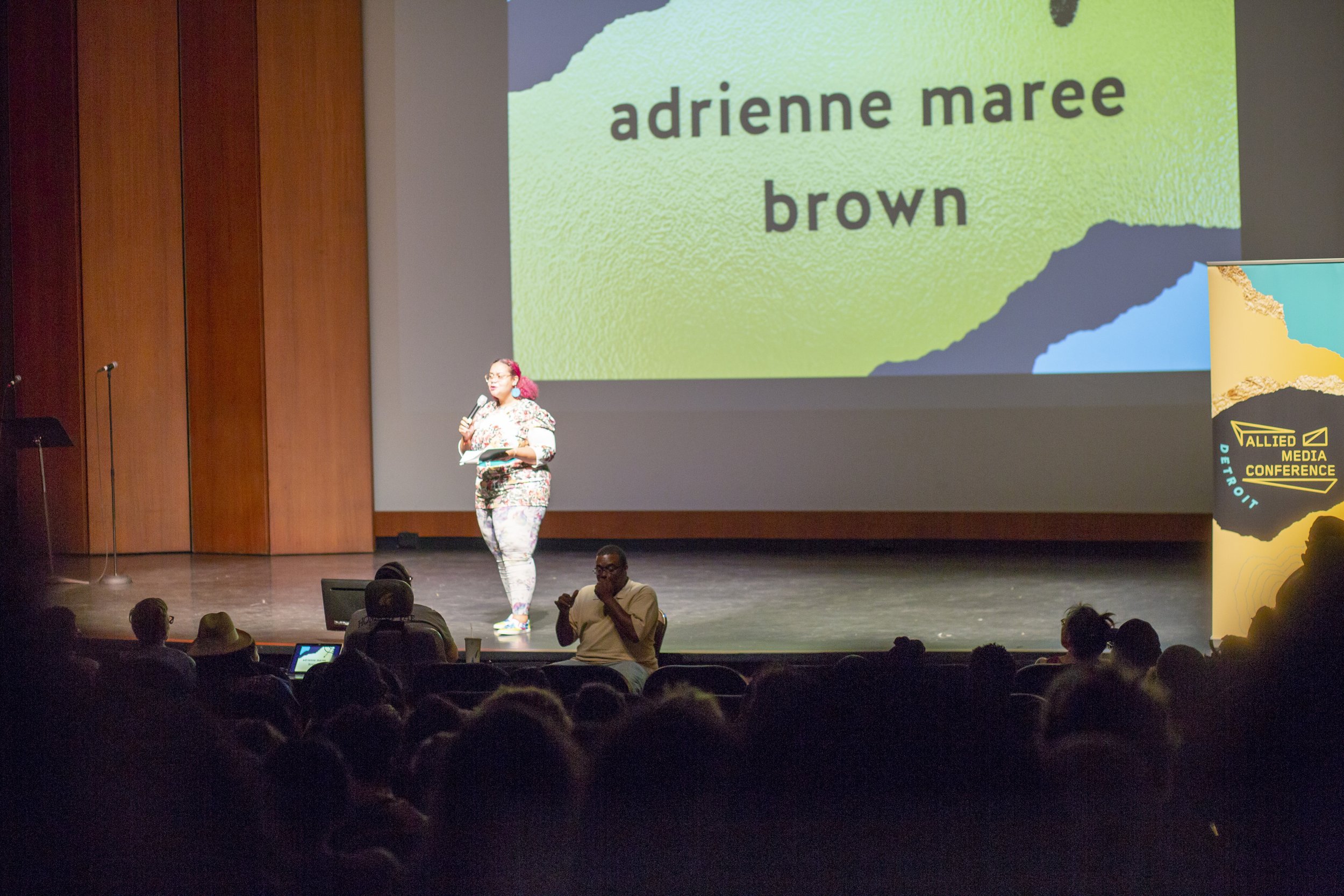 A woman on stage at a media conference speaking into a microphone, with a large screen behind her displaying the words 'adrienne maree brown' and a sign on the side reading 'Allied Media Conference Detroit'. The audience is seated in front of the stage.