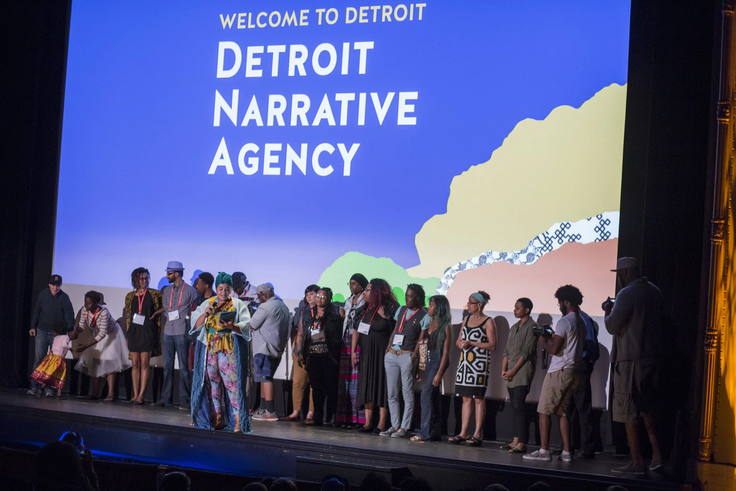A group of diverse people standing on stage in front of a large screen that says 'Welcome to Detroit, Detroit Narrative Agency'.
