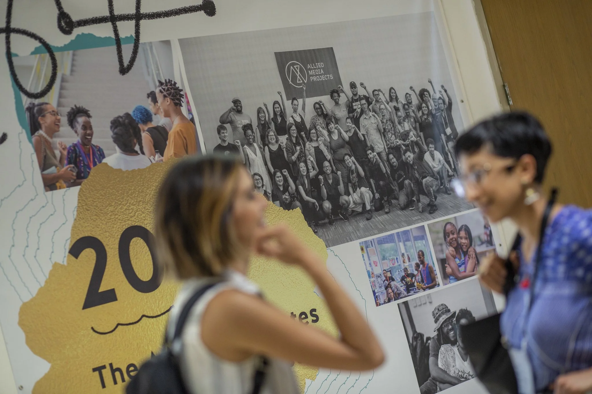 People attending a conference or event, with a poster on the wall showing photographs of diverse groups and activities, including a group photo with a sign 'Allied Media Projects' and smaller images of people engaging in various activities.