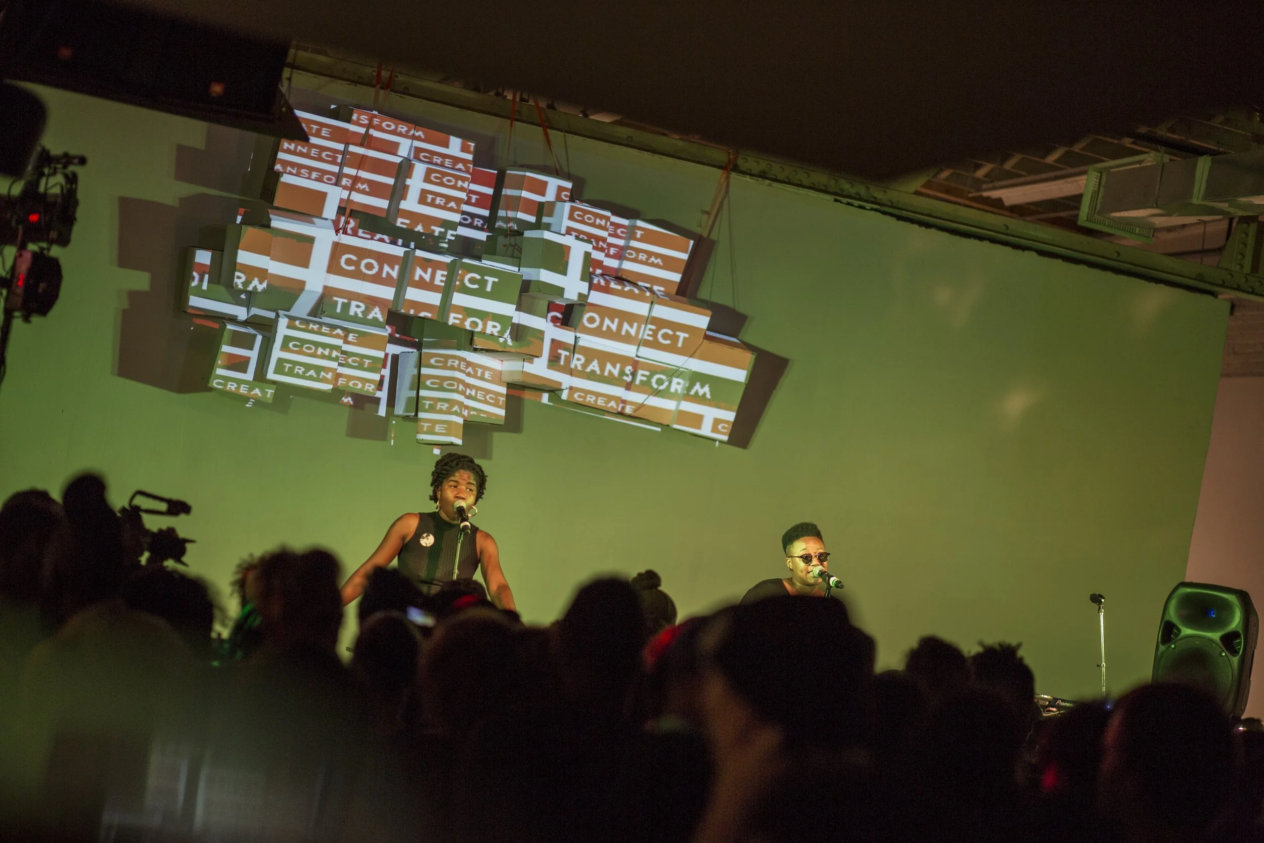 A live performance on stage with two singers, a woman on the left and a man on the right, both singing into microphones. The stage has a green background with a large hanging display of screens showing the words "CONNECT," "CREATE," and "TRANSFORM." There is an audience in the foreground.