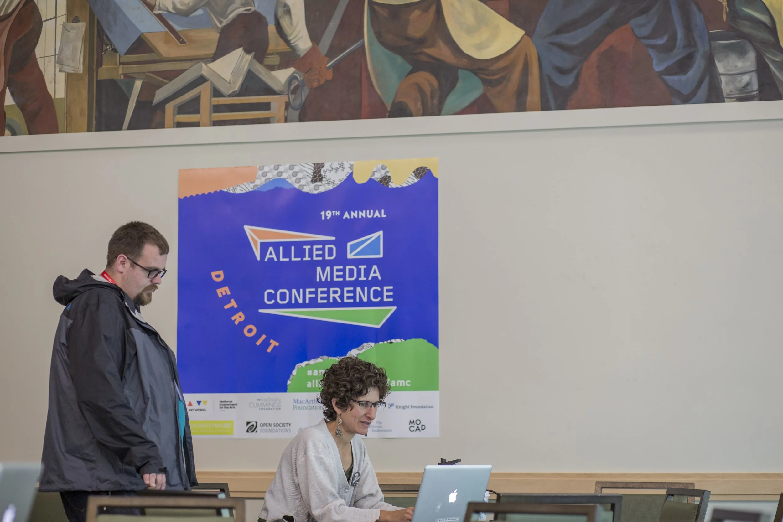 A man and a woman at a conference registration table, with a large blue and green conference poster behind them.