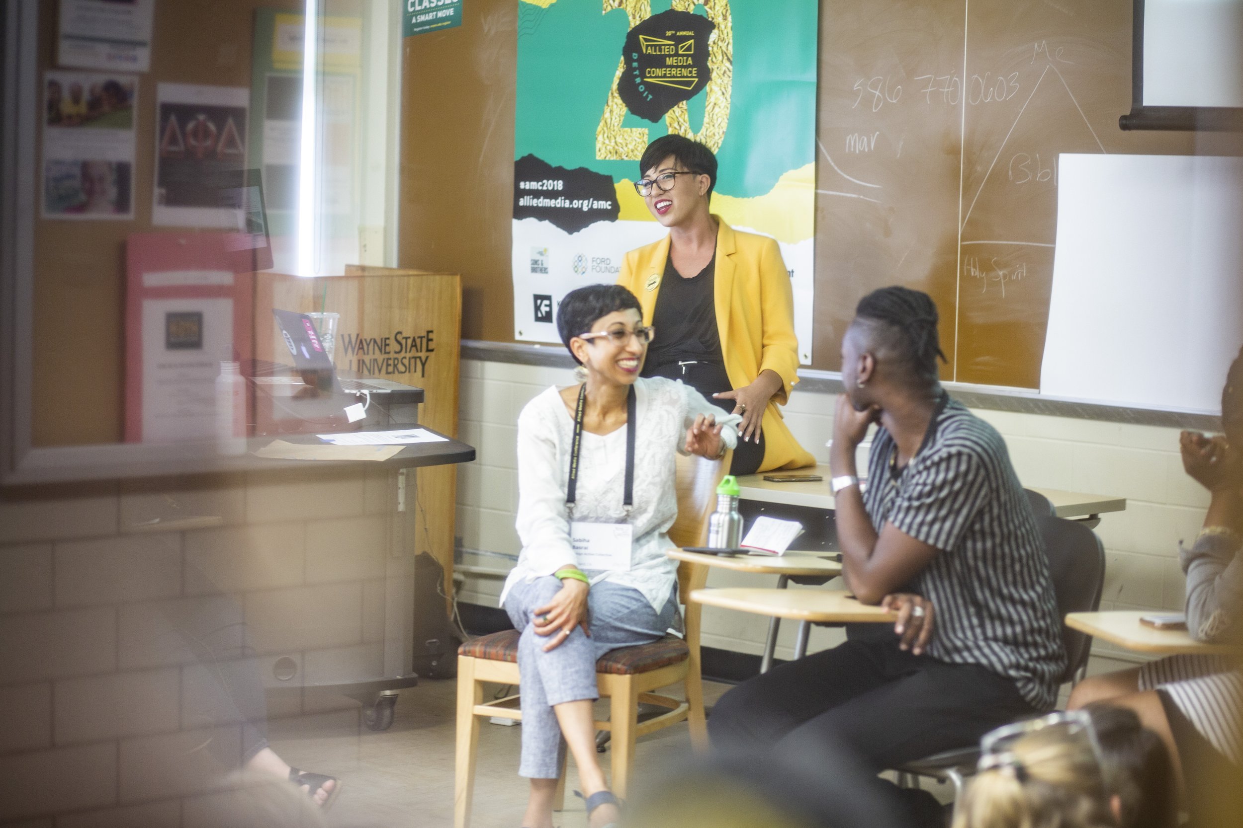 A classroom or conference room setting with three women engaging in conversation. One woman with short hair and glasses sitting on a chair is gesturing while speaking. Another woman with glasses and a striped shirt is sitting at a desk, listening attentively. A third woman with short dark hair and wearing a yellow blazer is standing at the front near a blackboard, smiling. There is a poster for an media conference on the wall behind her, and the event appears to be at Wayne State University.