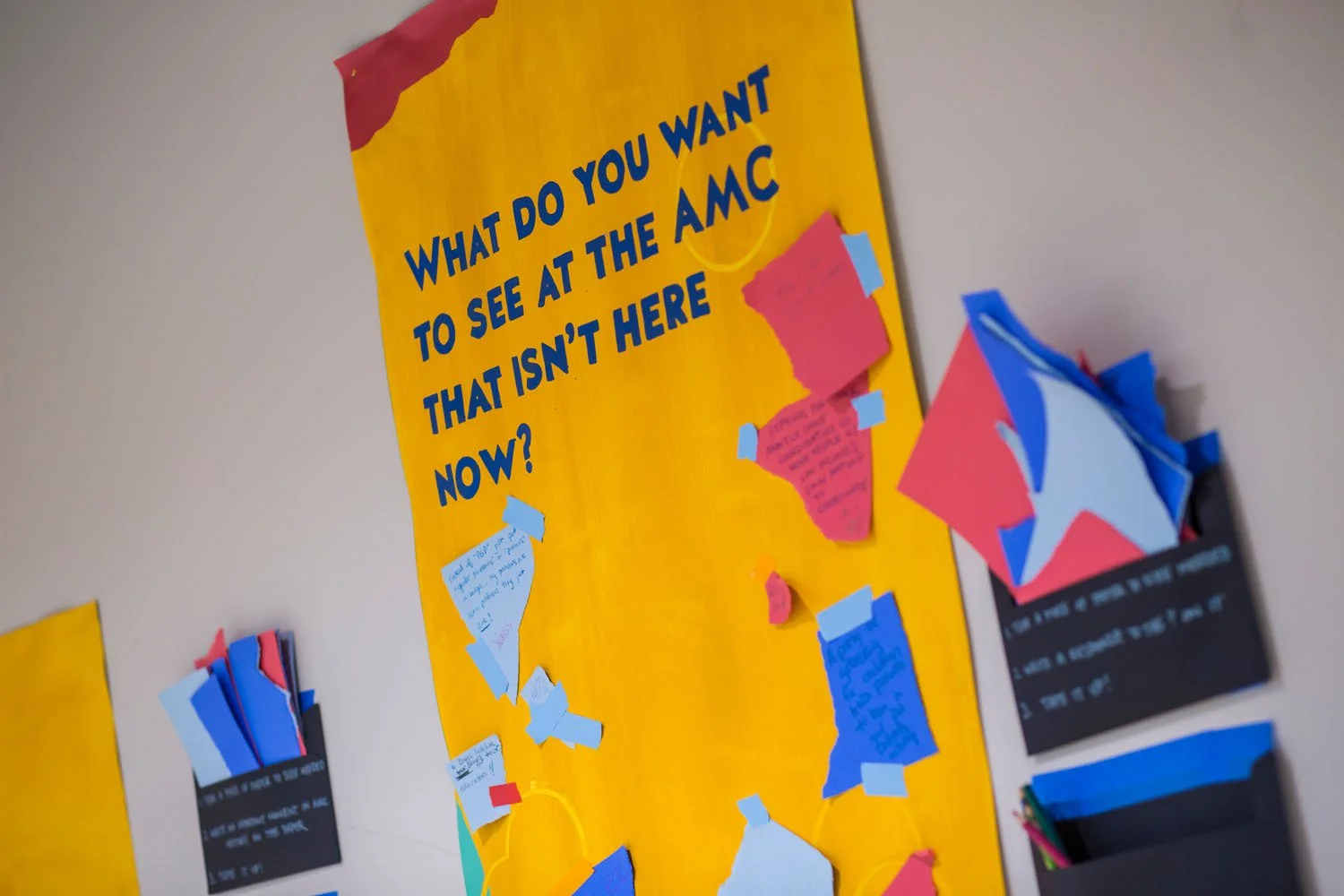 Bulletin board with yellow background and blue text asking, "What do you want to see at the AMC that isn't here now?" There are pink, blue, and other colored paper notes taped to the board and other note cards on black pocket organizers attached to the wall.