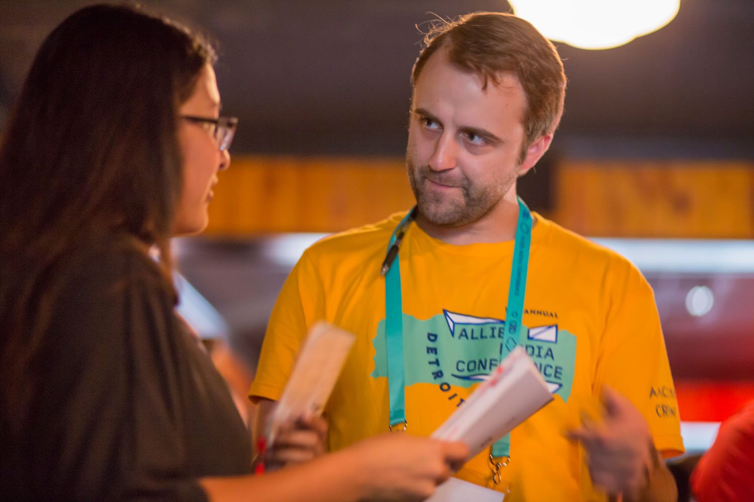 A man in a yellow conference T-shirt with a blue lanyard conversations with a woman holding a notepad.
