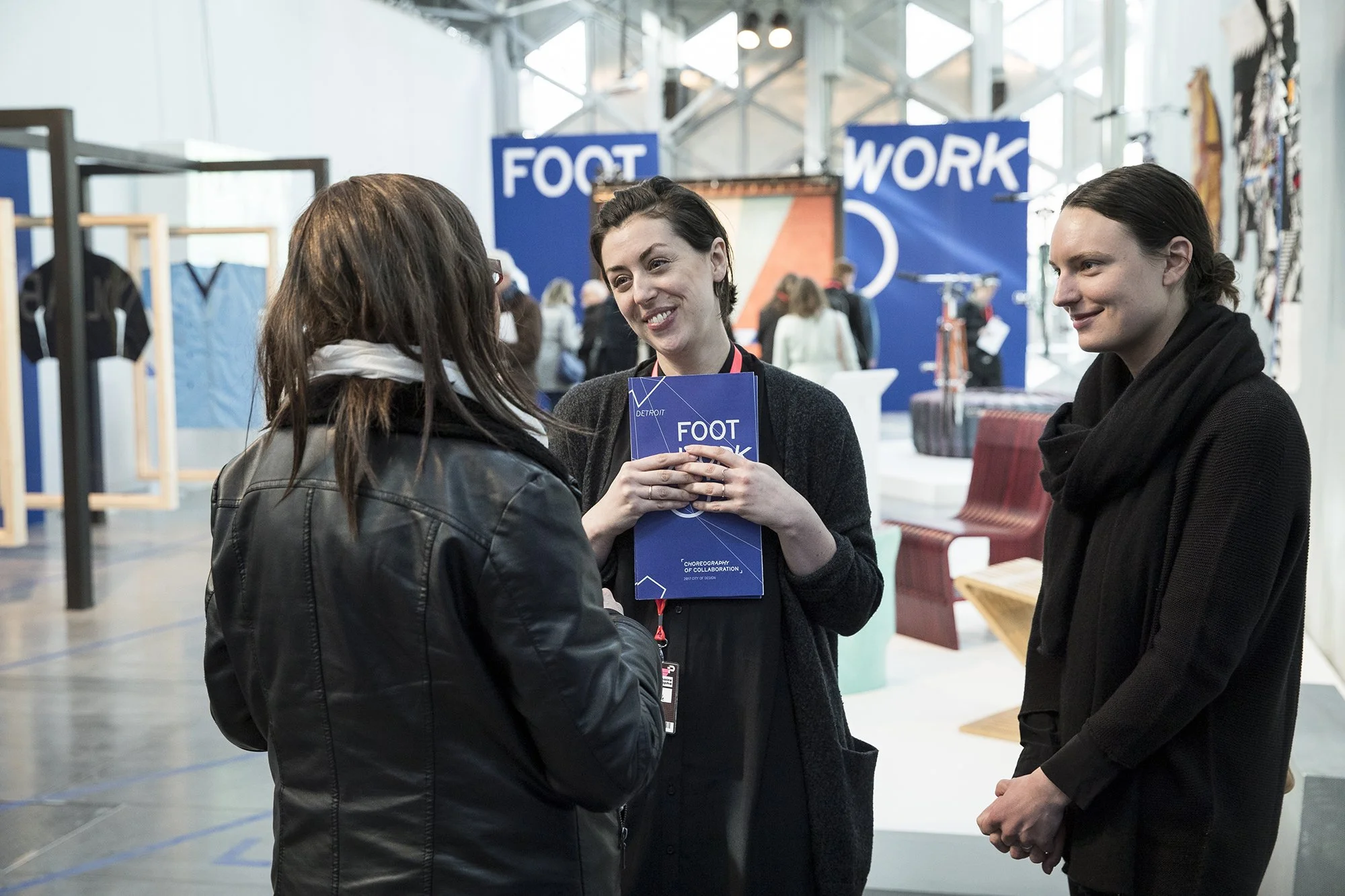 Three women engaging in conversation at an indoor event, with signs in the background that read 'FOOT' and 'WORK'.