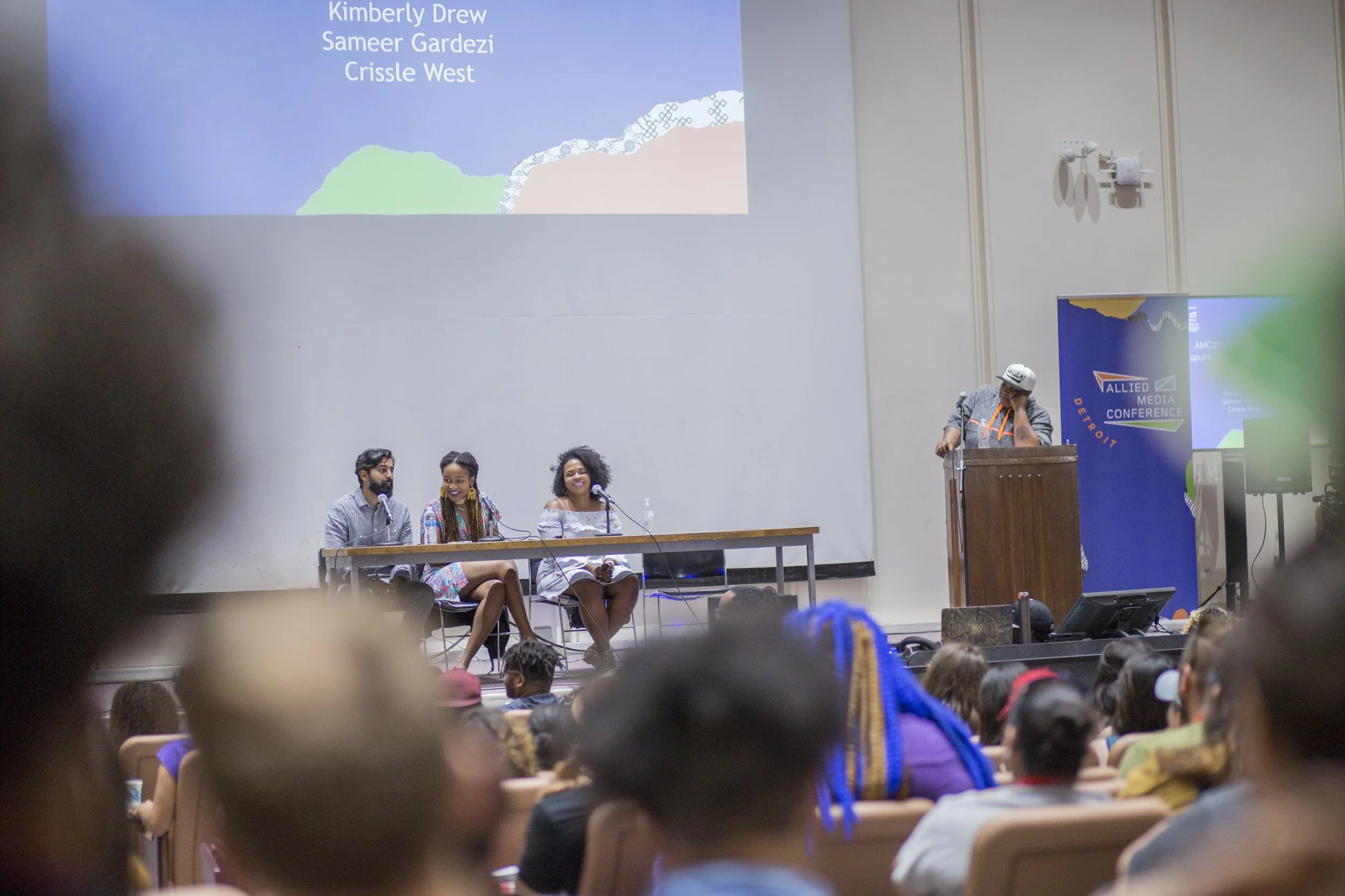 Panel discussion on stage with four people, three women and one man, sitting at a table with microphones, in front of an audience in a conference room. A large screen above displays names, and a sign on the right reads "Allied Media Conference."