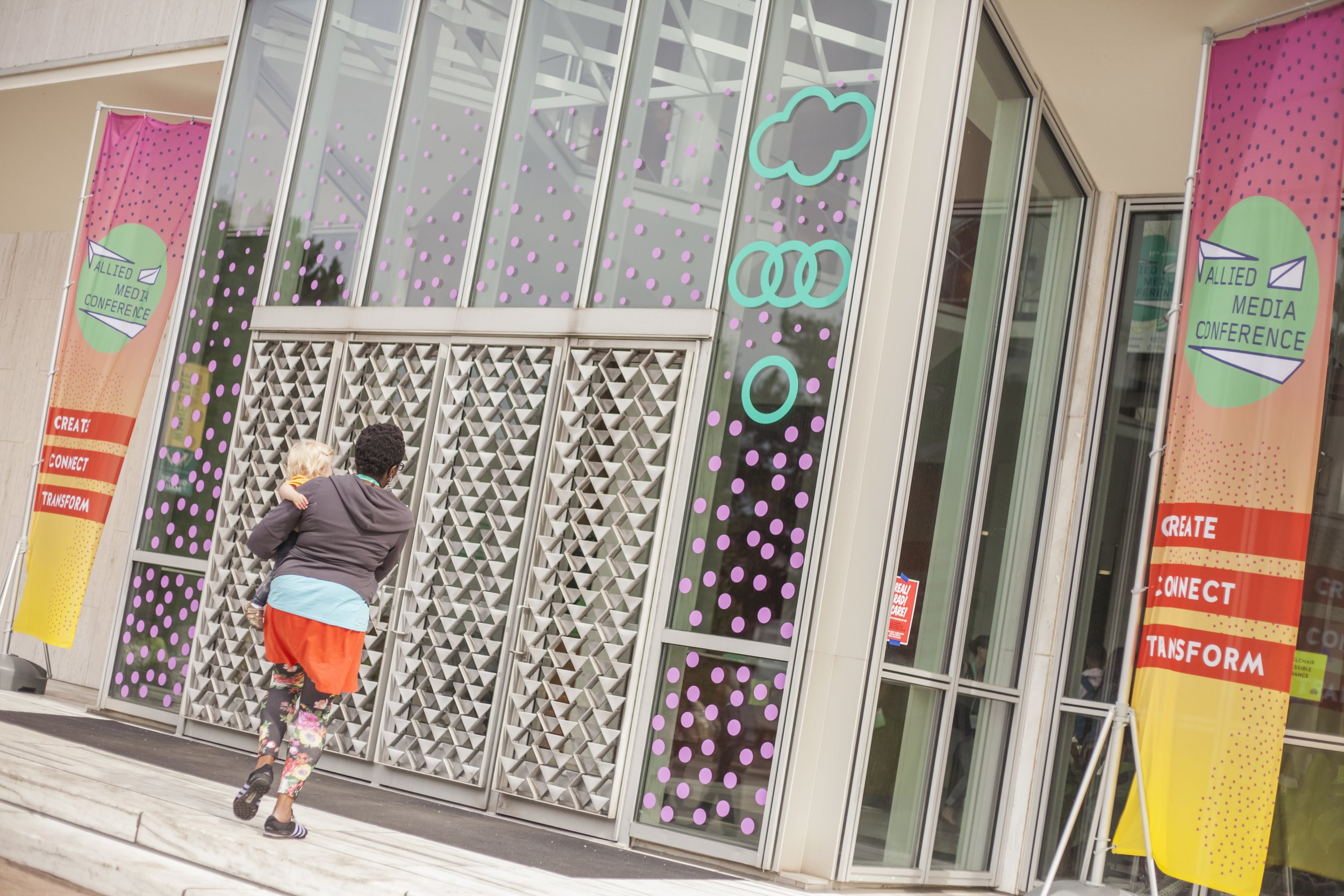 Person carrying a child into a conference center with banners that read 'Allied Media Conference' and 'Create, Connect, Transform'