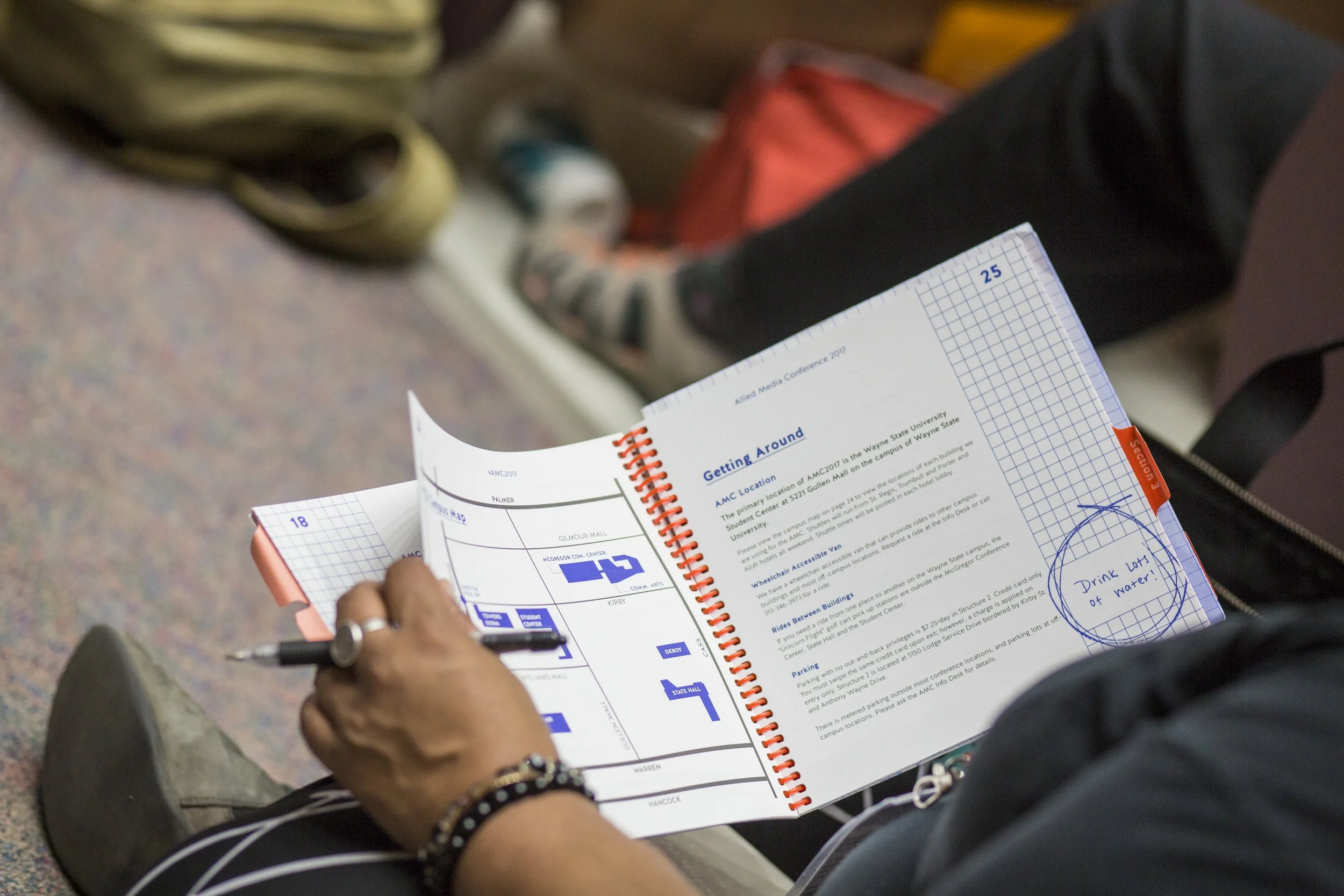 Person holding a conference guide and pen, sitting on the floor at an event with other attendees, with a backpack nearby.