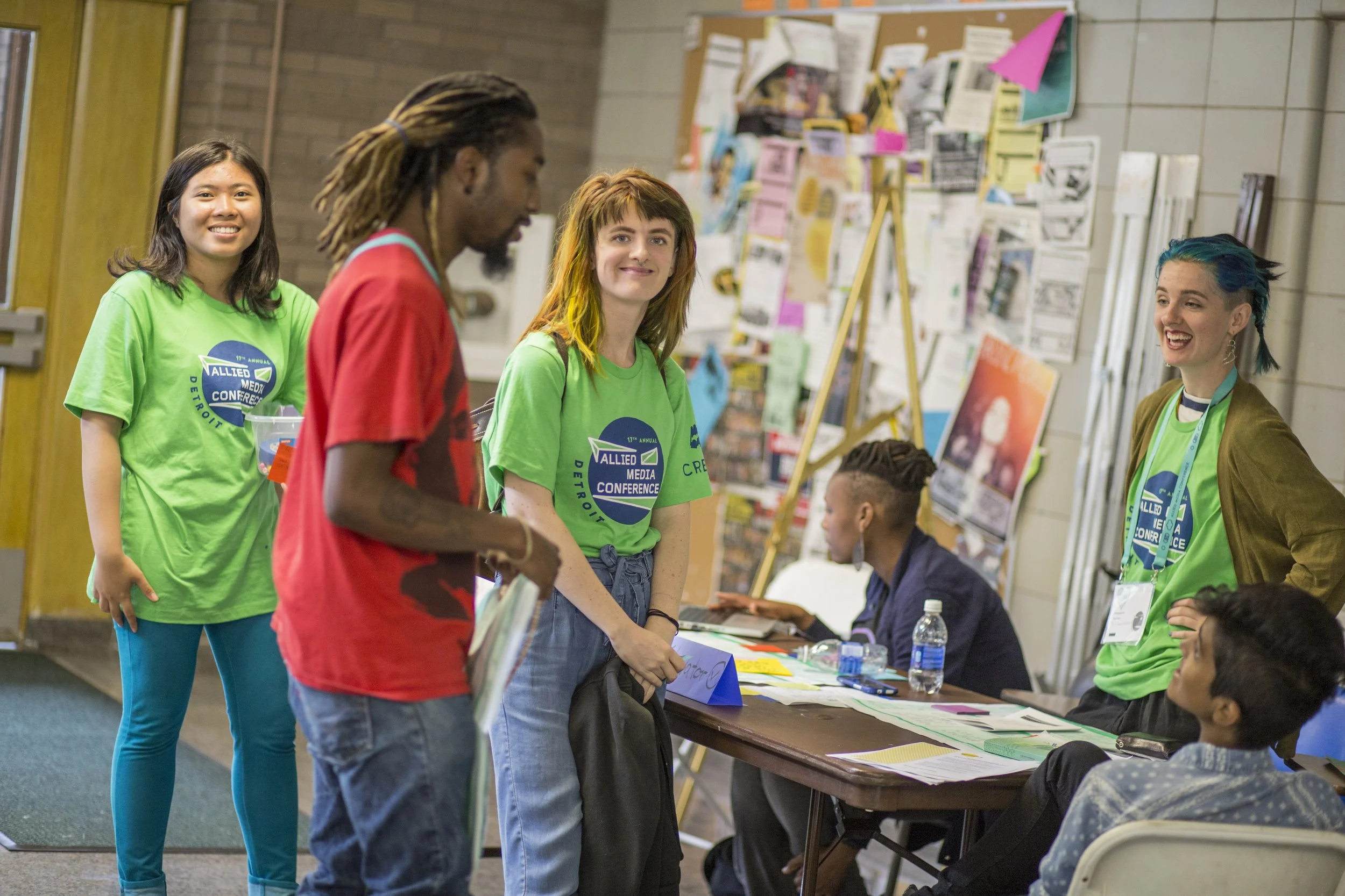 A group of five diverse young adults at a registration or information desk at a conference, with a woman in a blue shirt sitting behind the table and a colorful bulletin board in the background.