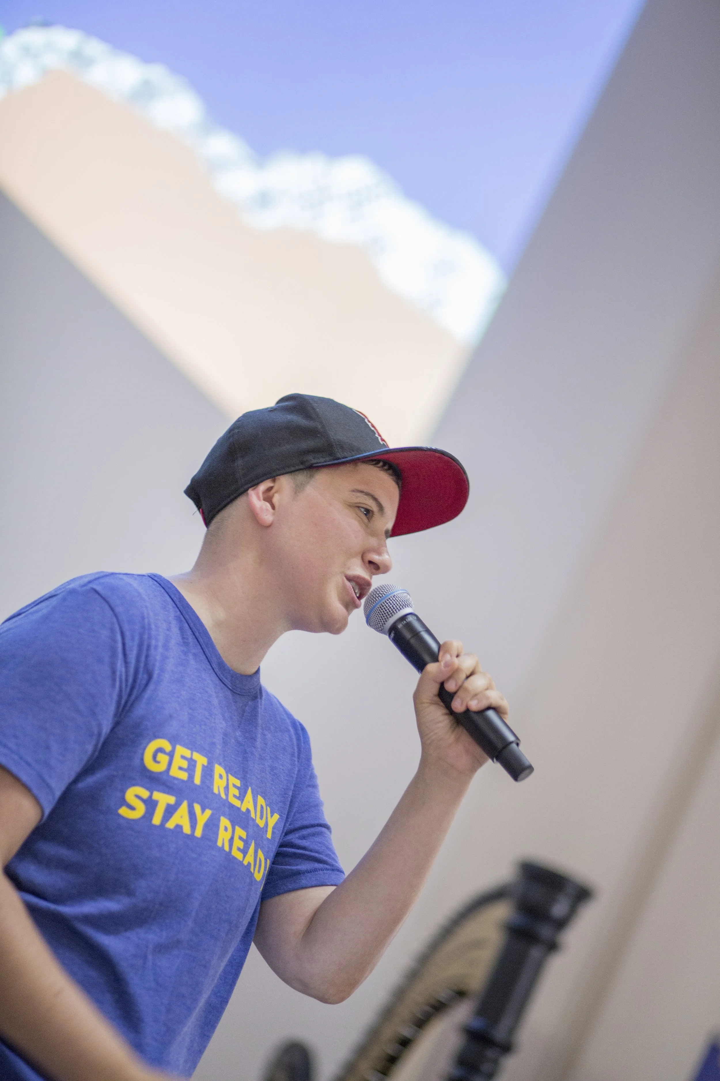 A person holding a microphone, speaking or singing, wearing a blue t-shirt with yellow text that reads 'GET READY STAY READY' and a black baseball cap with a red brim.