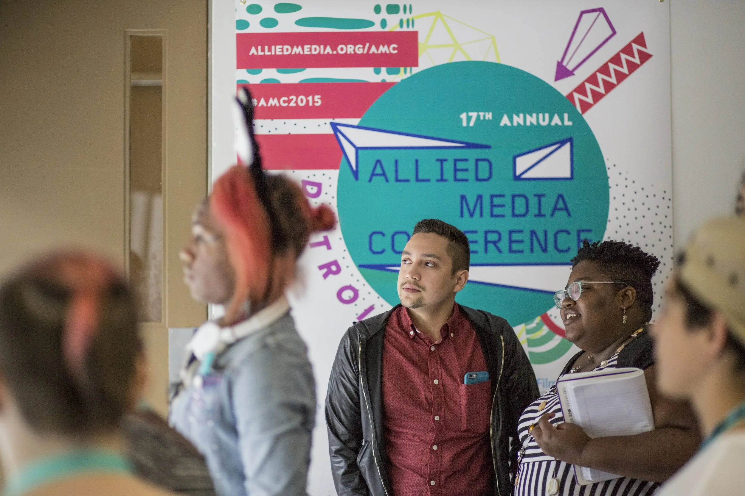 People attending the 17th Annual Allied Media Conference. The conference poster behind them displays the URL alliemedia.org/amc and the hashtag #AMC2015.