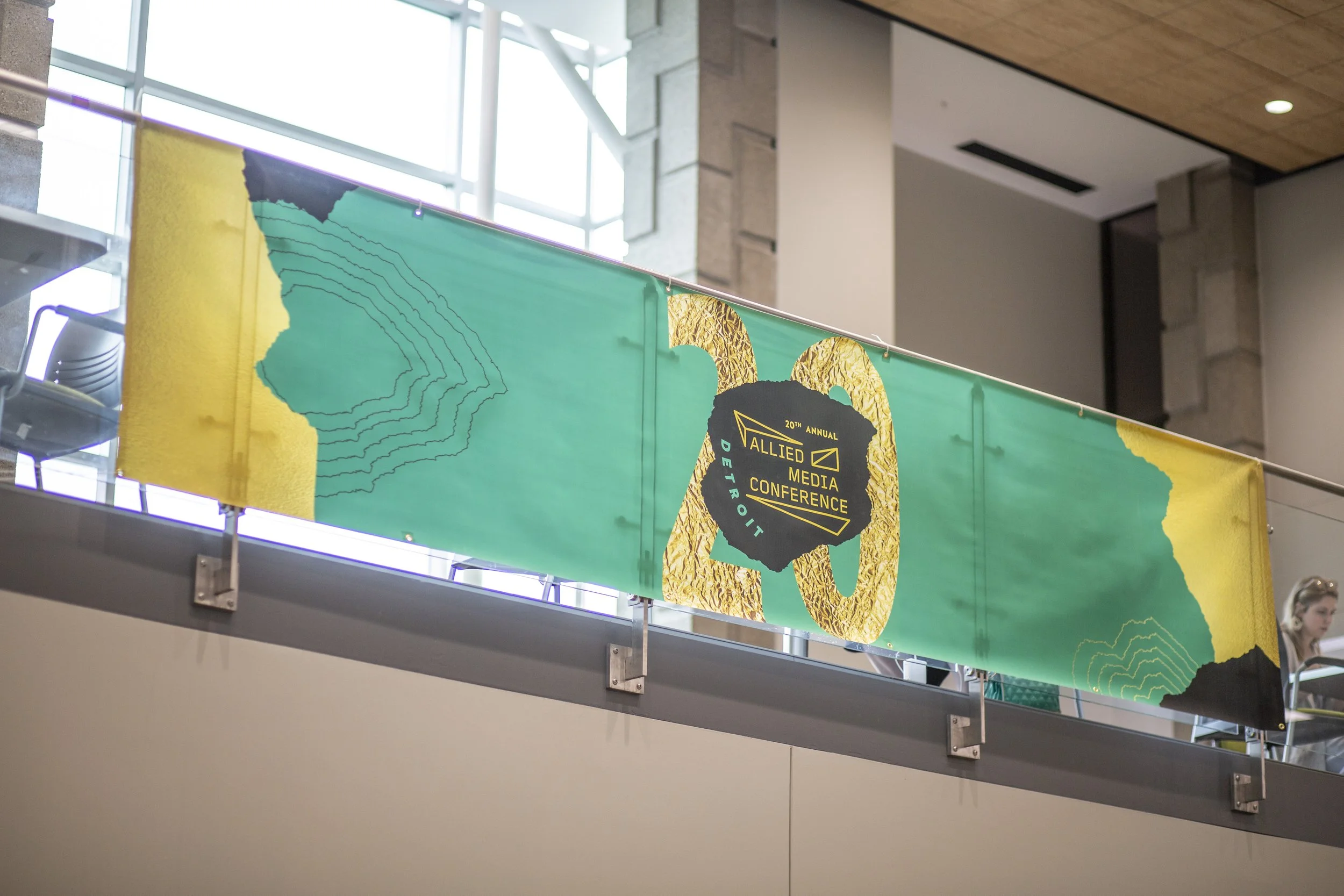 Conference banner hanging inside a modern building with large windows, featuring the logo and name of the 20th Annual Allied Media Conference in Detroit, with abstract yellow, teal, and black design elements.