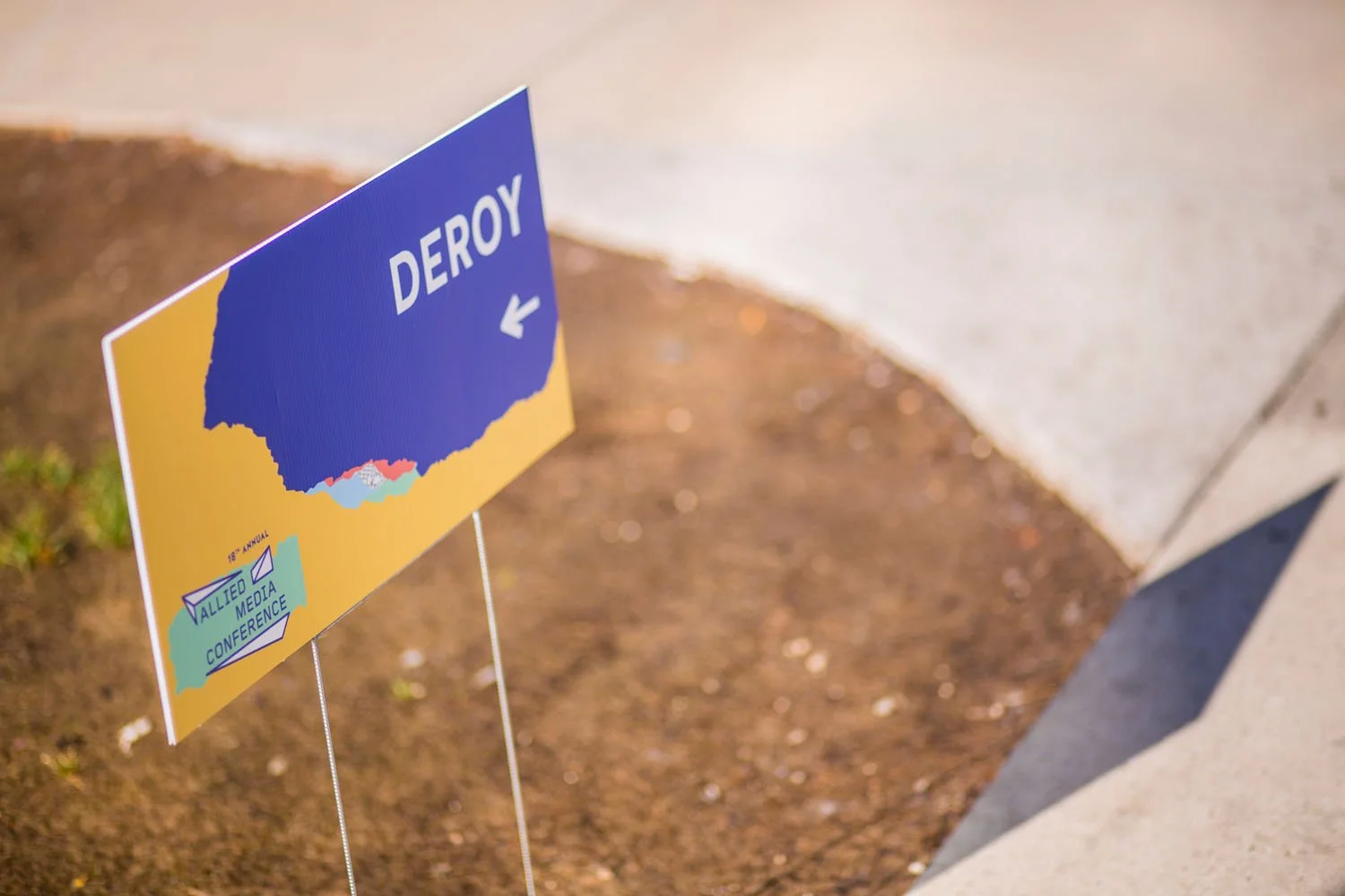 A political sign with a blue silhouette of a country and the word "DEROY" with an arrow pointing left, on a lawn next to a sidewalk.