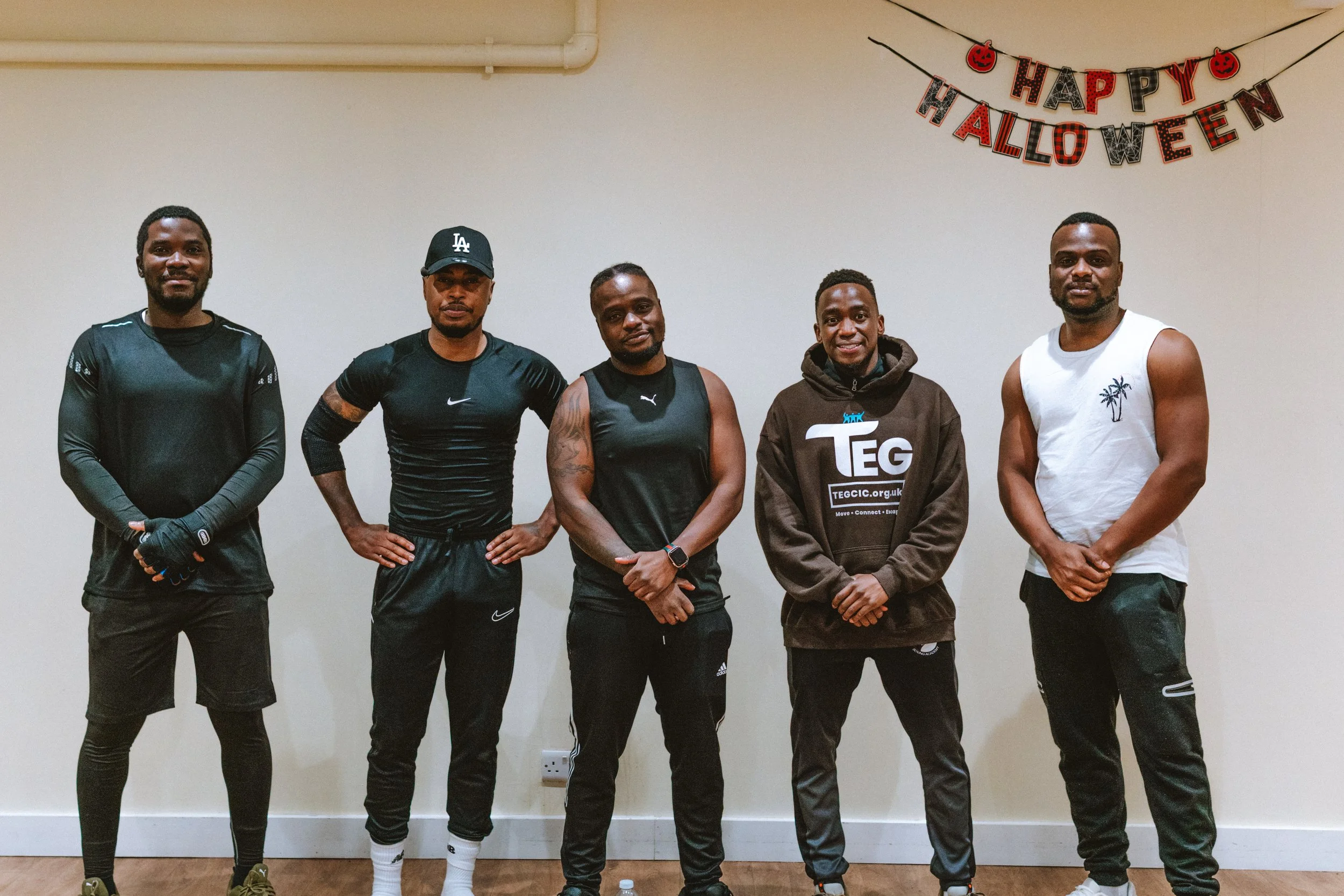 Group of five men standing indoors in front of a white wall with a Halloween banner that reads 'Happy Halloween' in red, black, and orange lettering, some with Halloween-themed decorations.