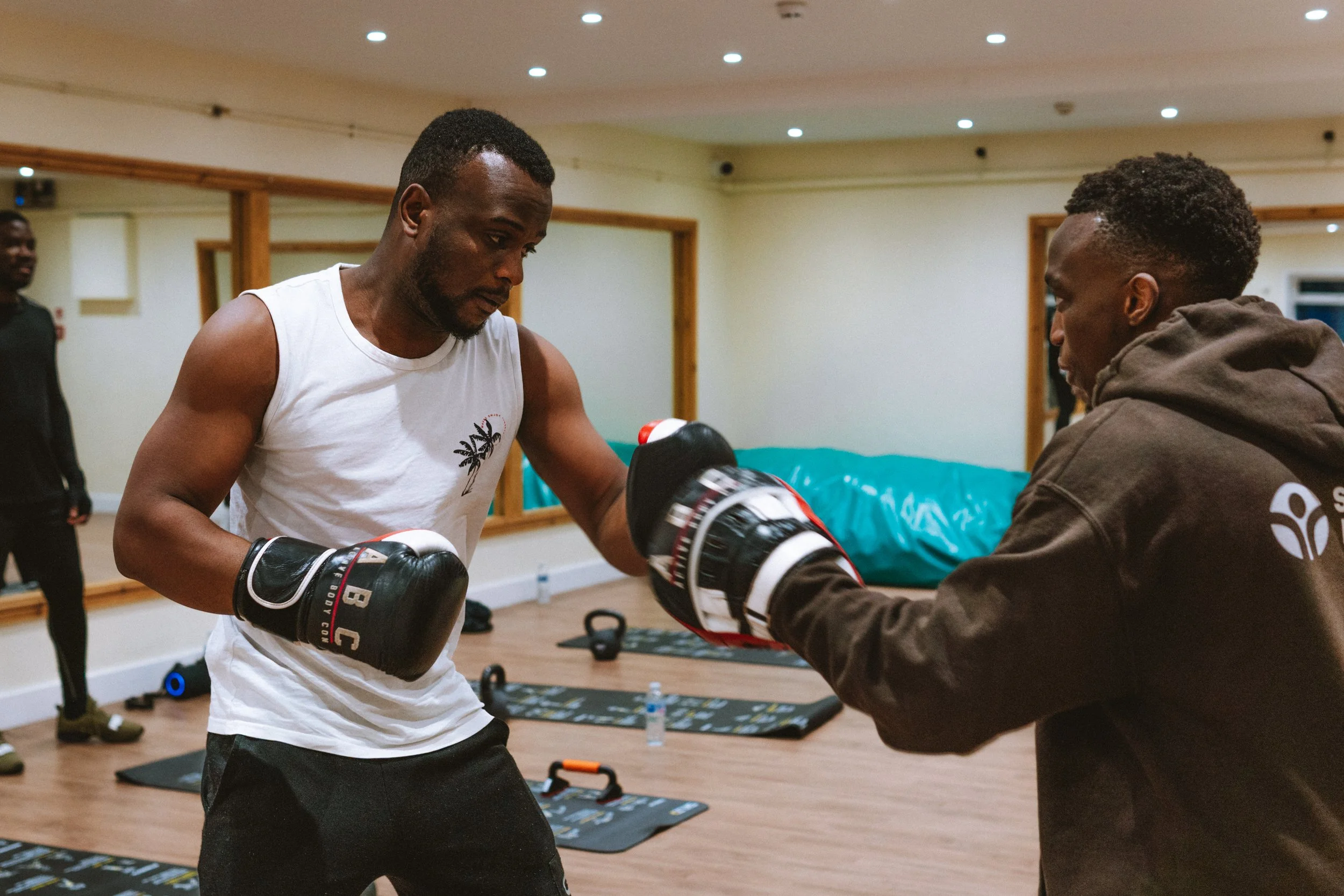 A man in a white tank top boxing gloves practicing boxing with a trainer in a gym. The trainer is holding focus mitts for him to punch. There are workout mats, kettlebells, and water bottles on the floor.