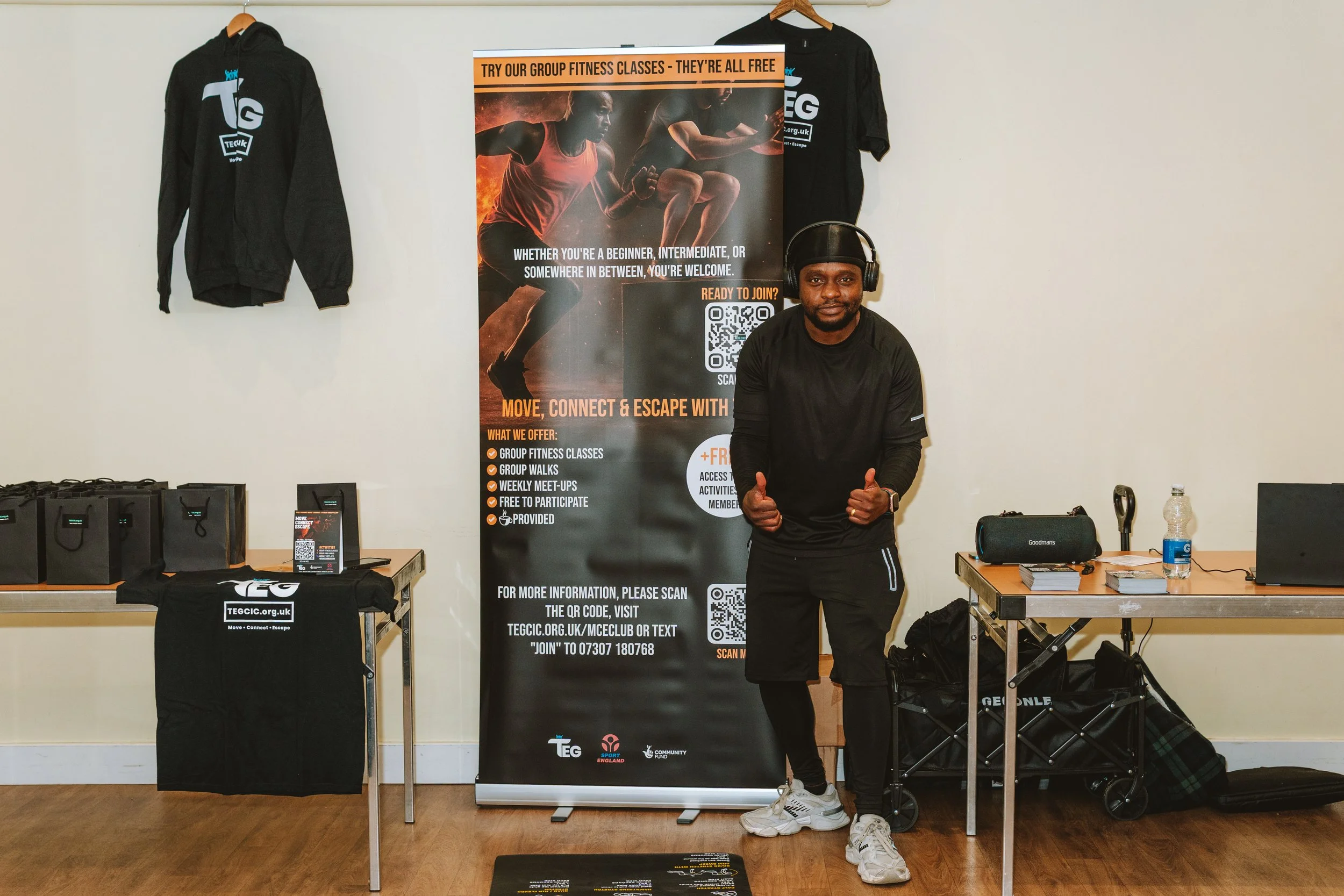 Man standing in front of a TE GCC promotional banner, giving a thumbs-up gesture. The banner advertises group fitness classes, walks, meet-ups, and other activities. A table on either side holds black bags, tablets, and a water bottle. Two black hood
