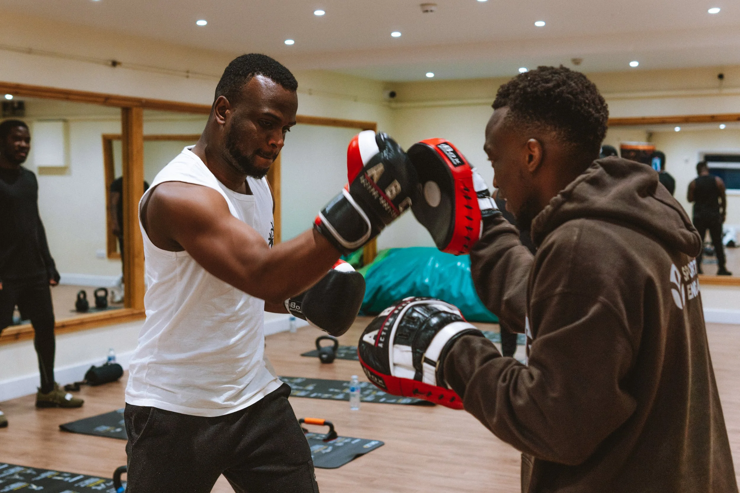 Two men practicing boxing with gloves in a gym, one wearing a white sleeveless shirt and the other a brown hoodie, while other people watch in the background.