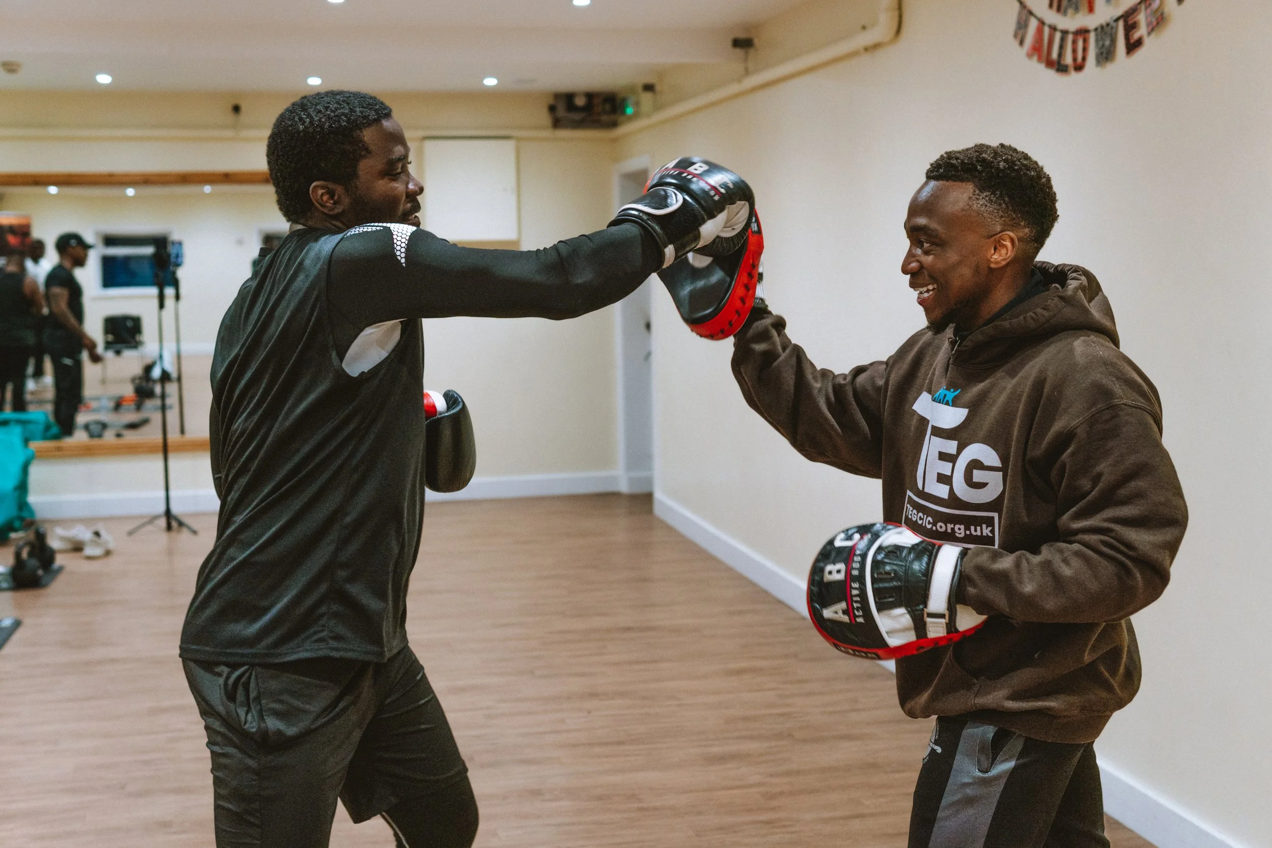 Two men practicing boxing with pads in a gym, smiling and facing each other.