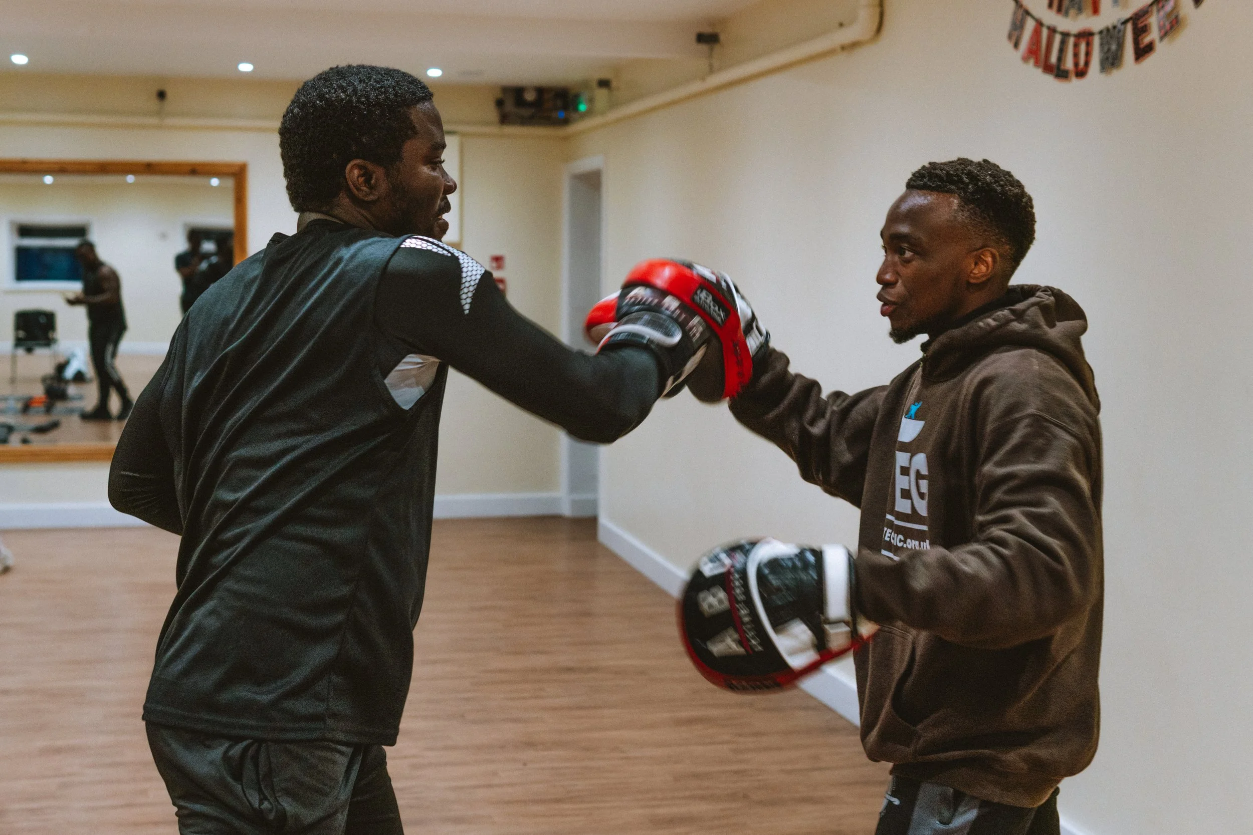 Two men in a gym engaging in boxing training; one is wearing boxing gloves and a sports shirt, while the other is holding boxing pads and wearing a hoodie.