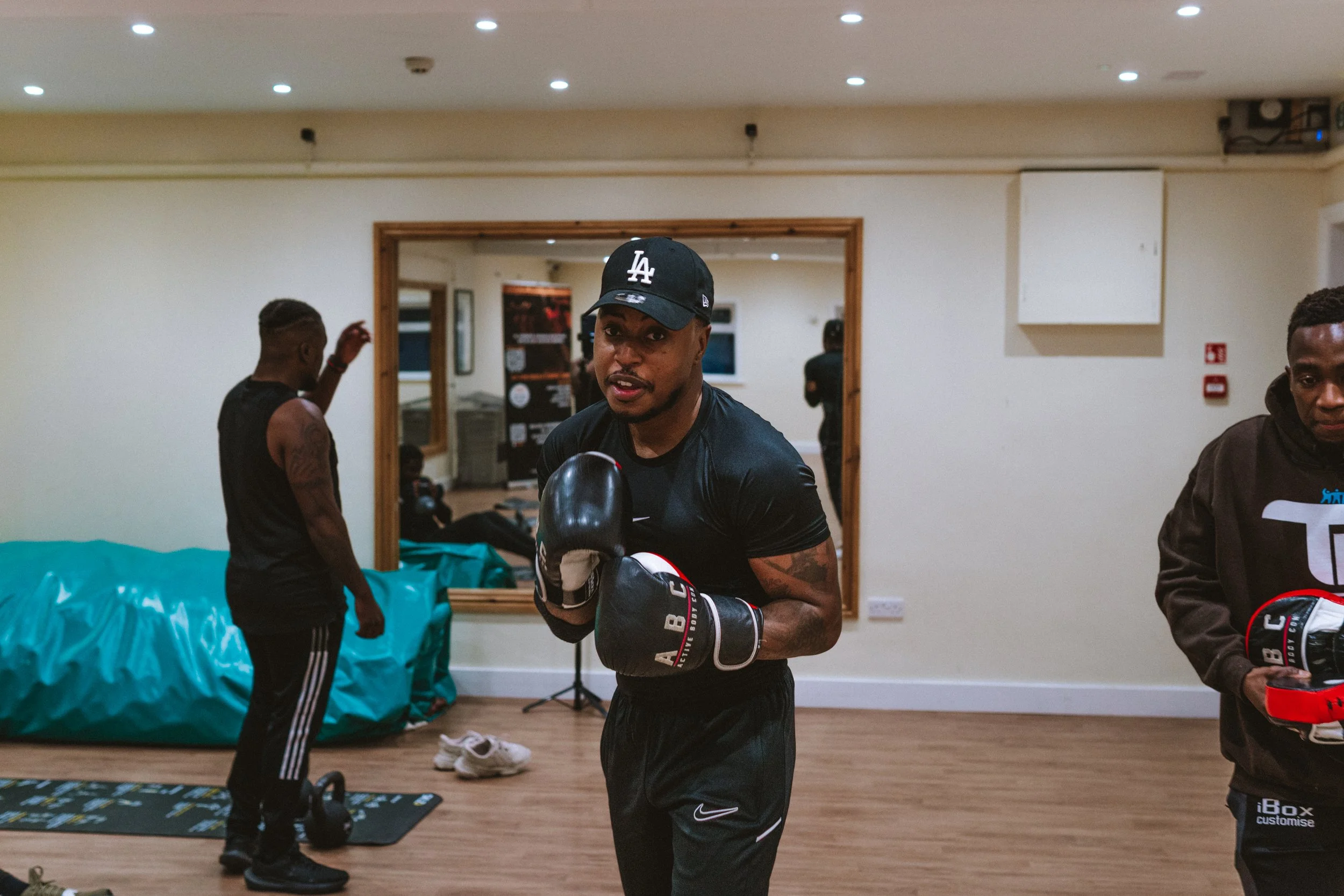 A man wearing a black Los Angeles Dodgers cap, black athletic shirt, and boxing gloves stands in a room, looking at the camera, with two others in the background, one adjusting his hair and the other holding boxing gloves.