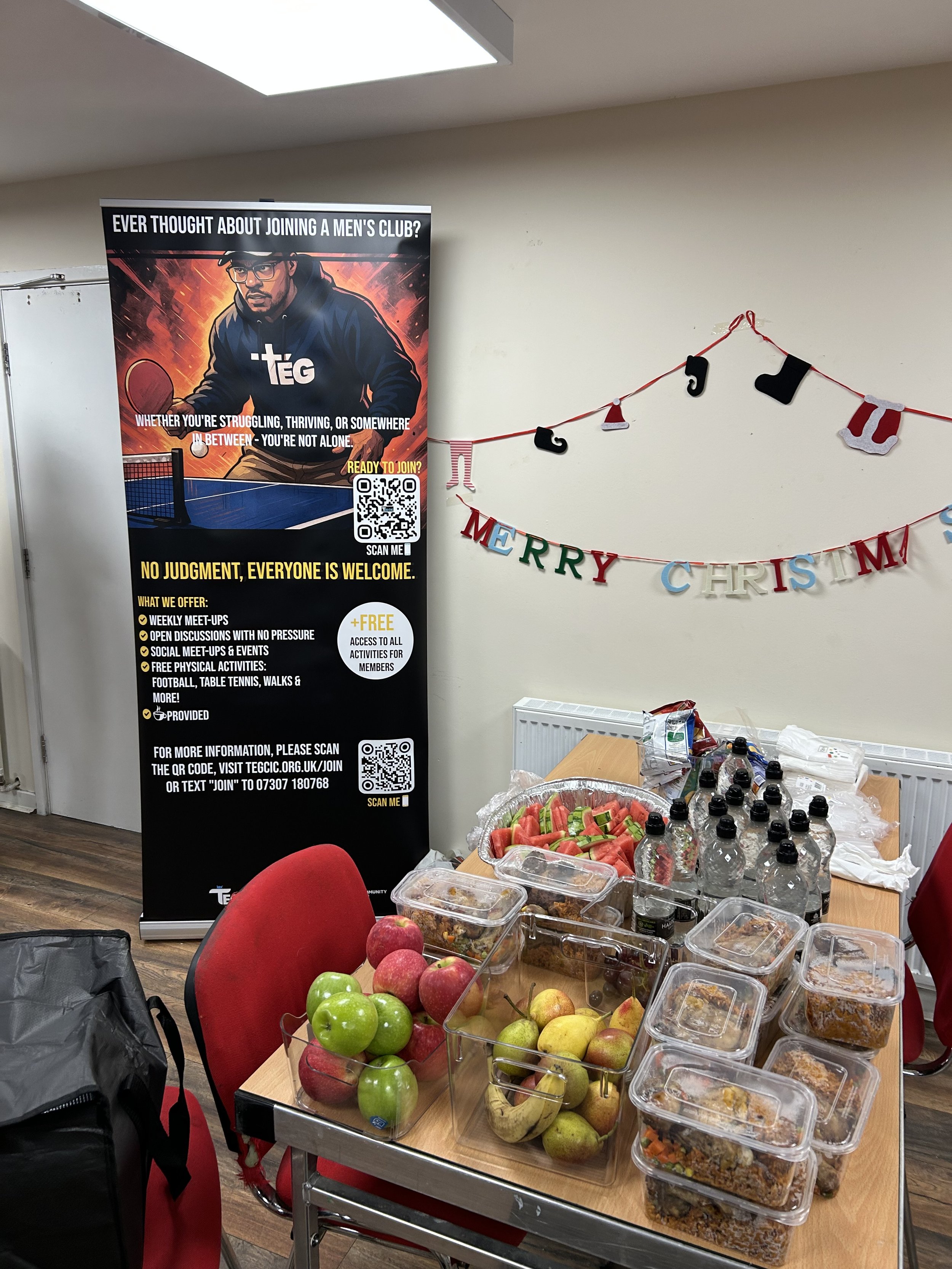 Table with apples, watermelon slices, bottled water, and food containers. In the background, a black and red banner for a men's club and a 'Merry Christmas' banner on the wall.