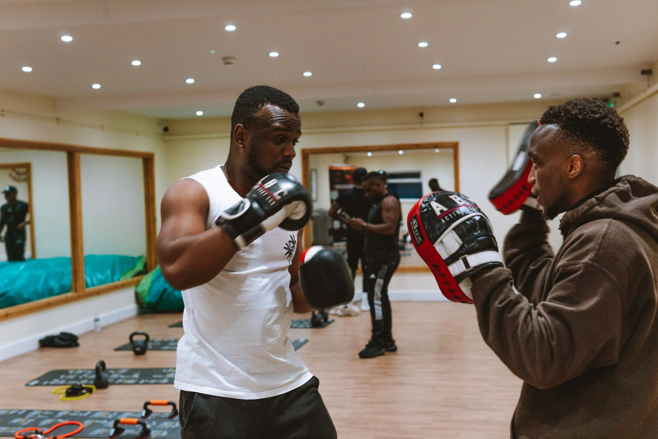 Two men practicing boxing in a gym, wearing gloves and holding focus mitts, with a mirror and workout equipment visible in the background.