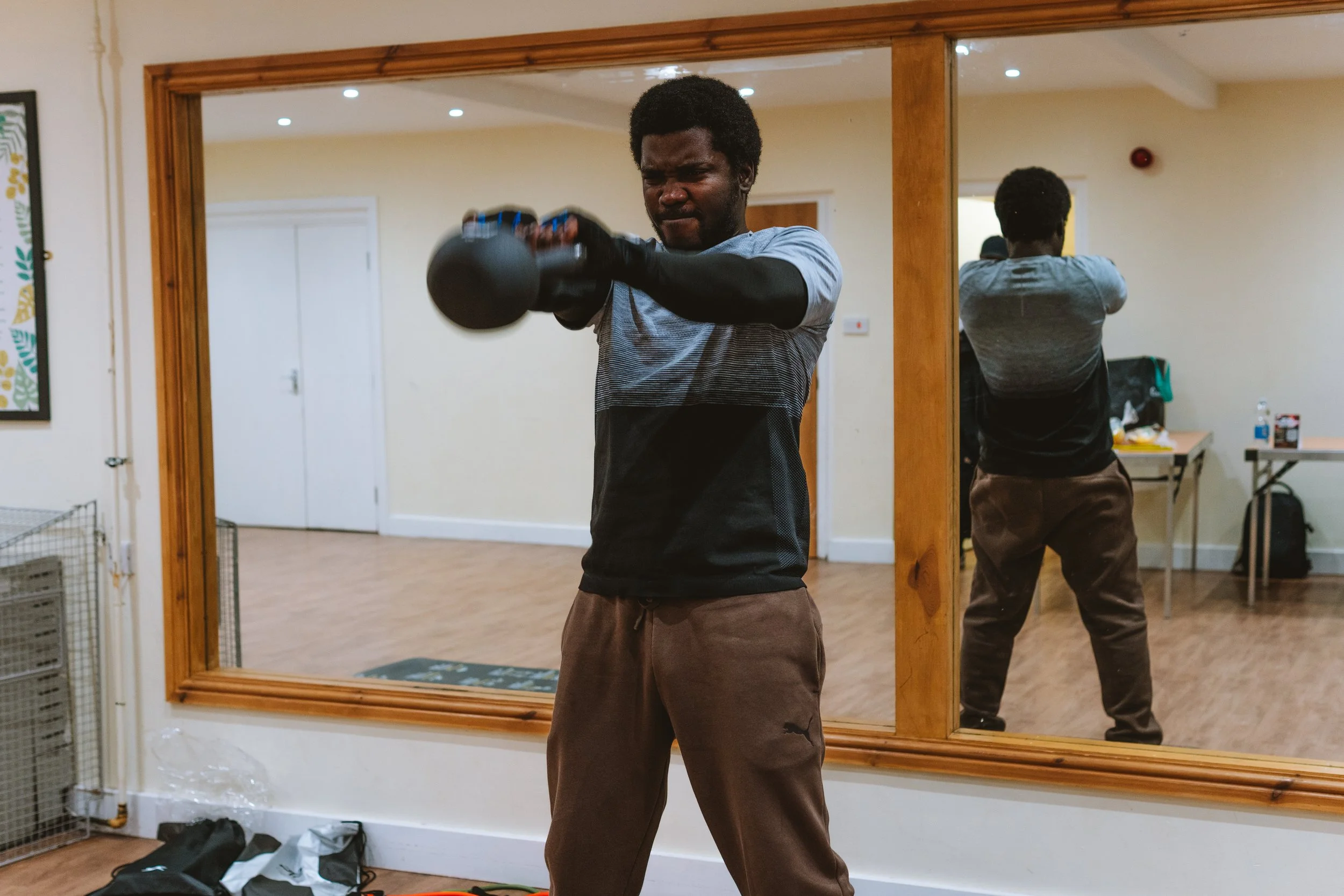 A man in gray and black workout clothes using a kettlebell in front of a large wall mirror.