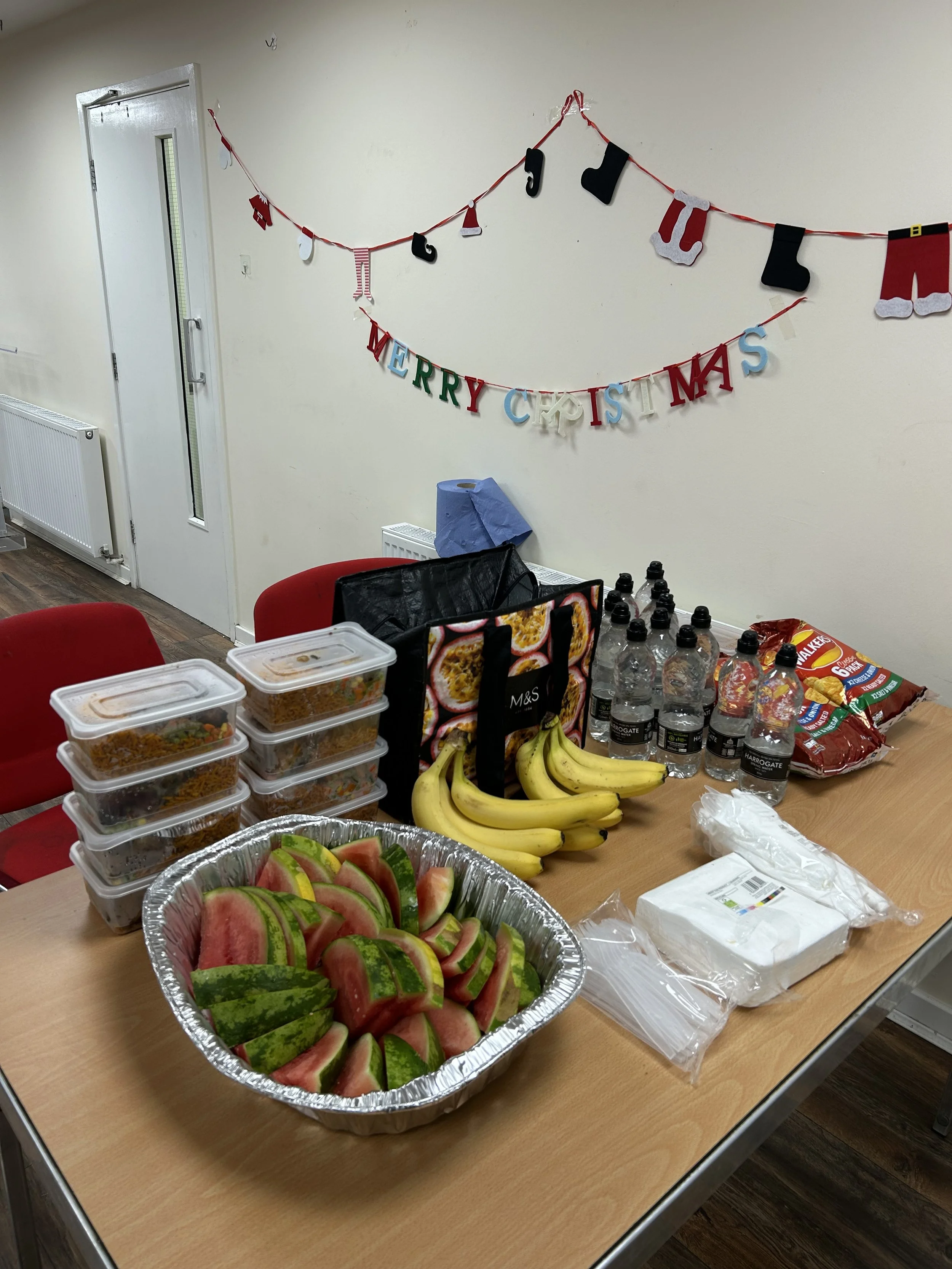 A table with stacked food containers, bananas, watermelon slices, bottled water, and snack bags, decorated with Christmas banner and stockings.