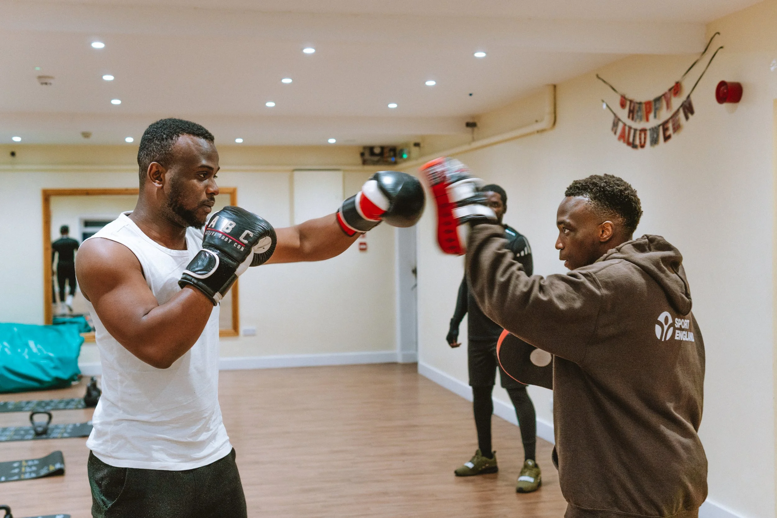 Two men practicing boxing with gloves in a gym, a trainer covering his face with his hands, and a workout room with a Halloween decoration on the wall.