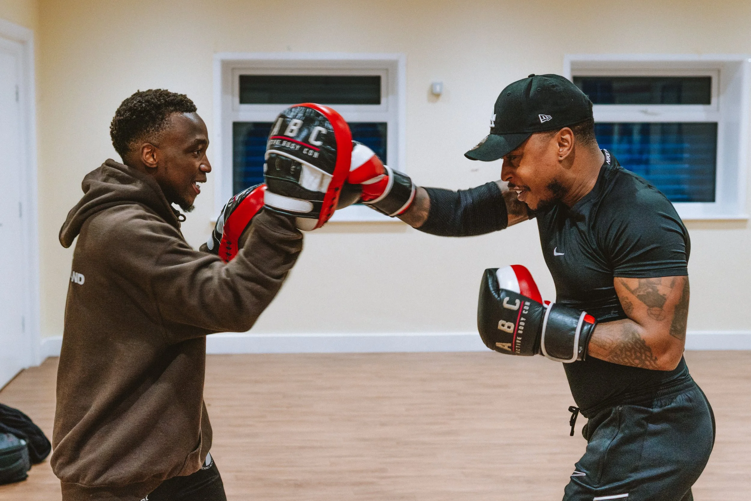 Two men engaged in boxing training, smiling and wearing boxing gloves, in a room with light-colored walls and wooden floors.