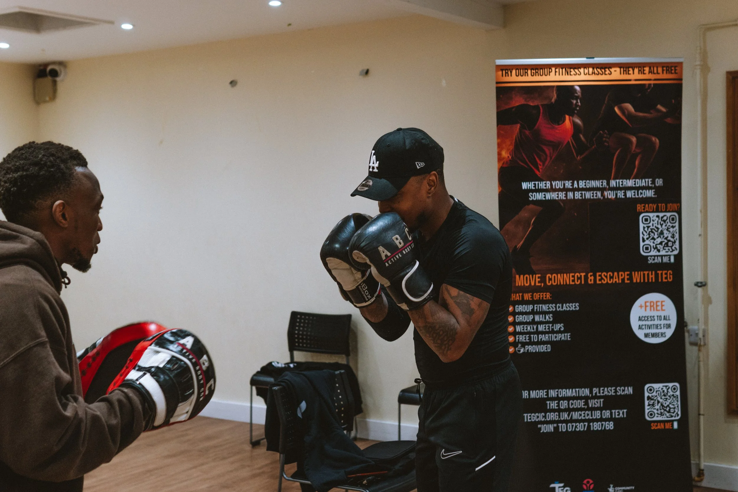 Two men in a boxing training session; one man is wearing boxing gloves and a cap, and the other is holding focus mitts. They are in a room with a fitness poster in the background promoting group fitness classes.