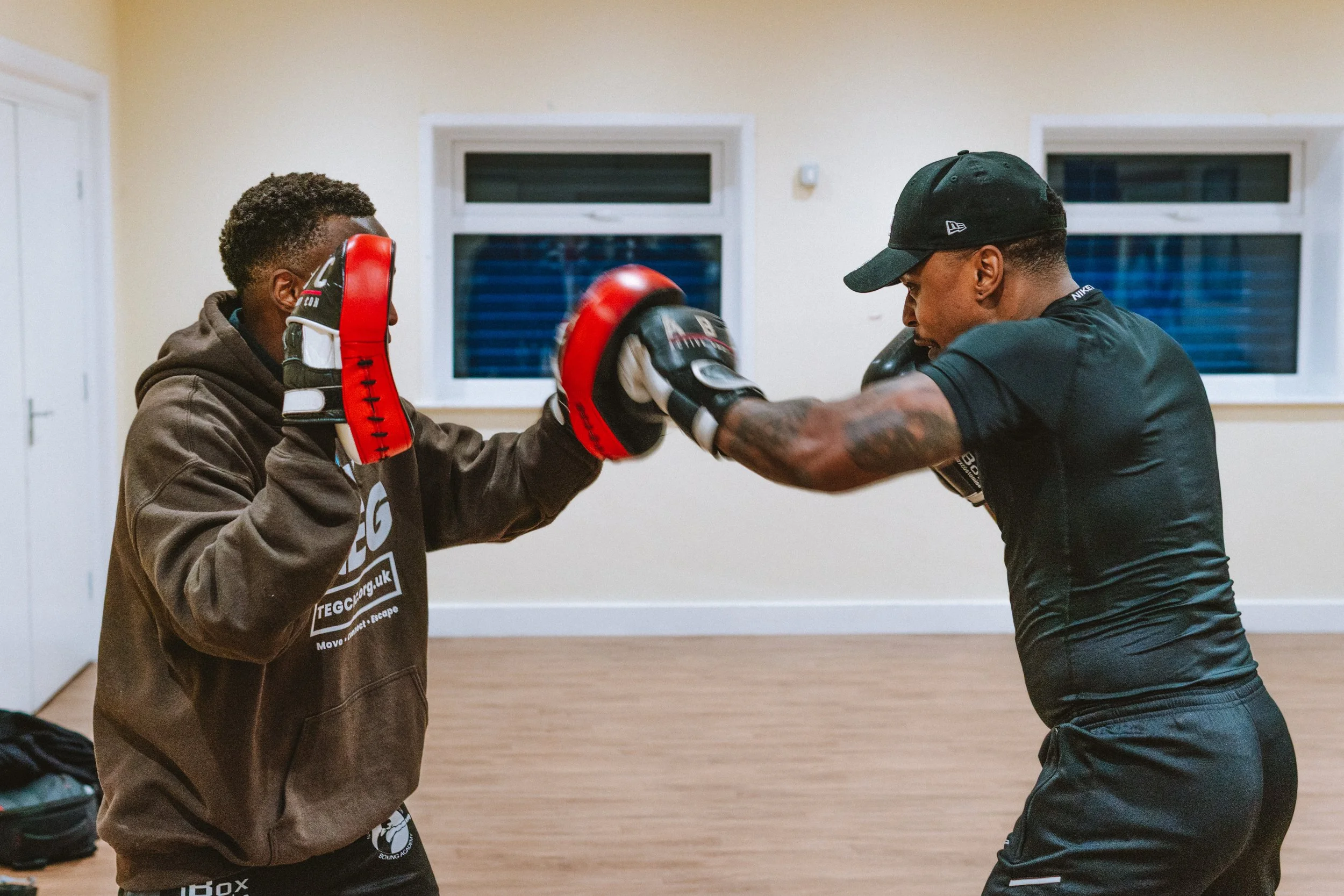 Two men practicing boxing with focus mitts in a room with wooden flooring and white walls.