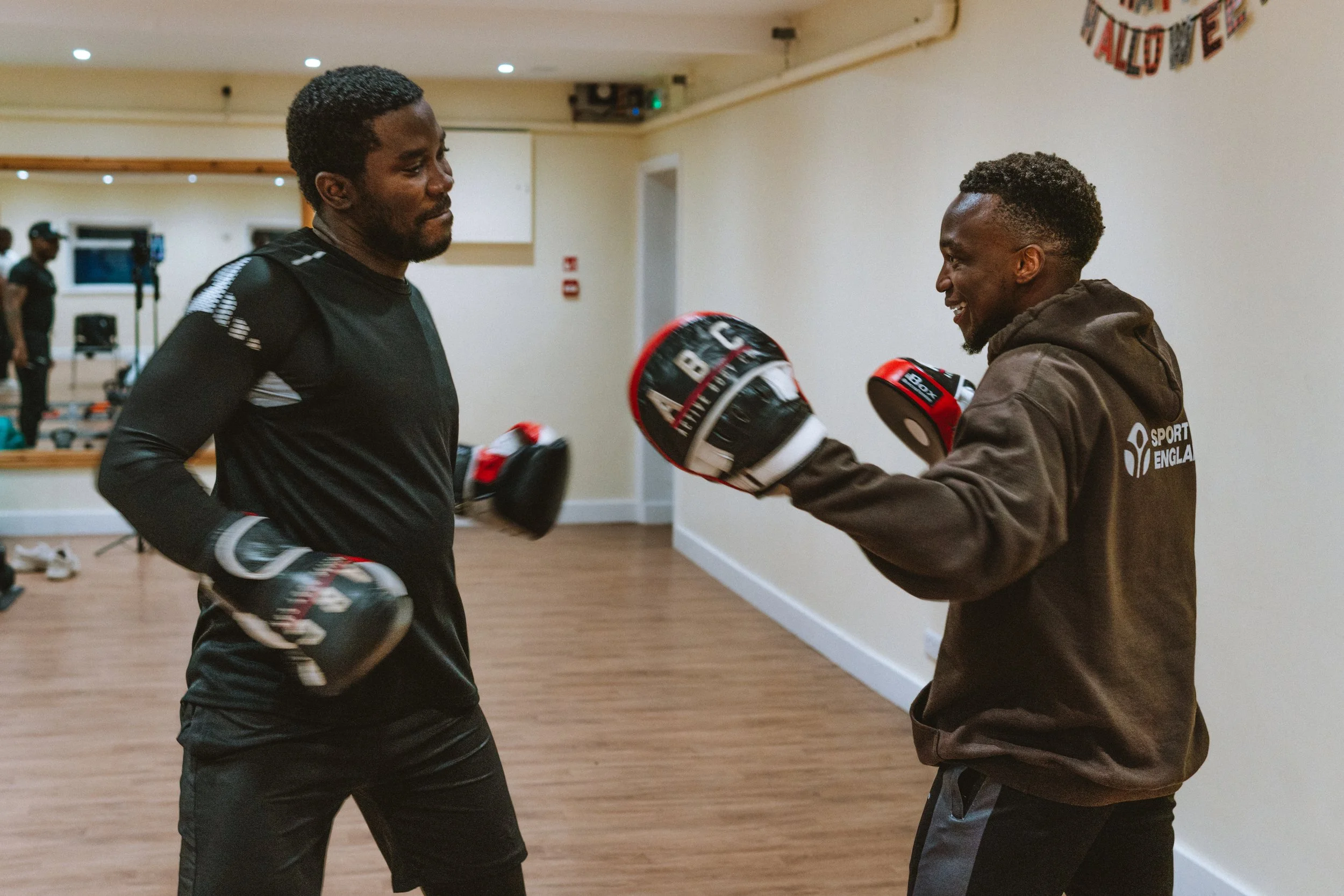 Two men training with boxing gloves and pads in a gym, smiling at each other.