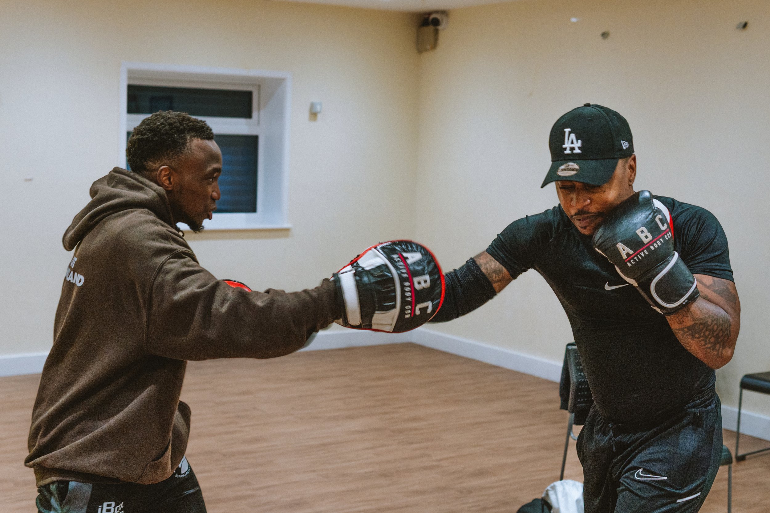 Two men practicing boxing in a training room. One man is throwing a punch while the other is blocking with his glove. Both are wearing boxing gloves and casual workout clothes.