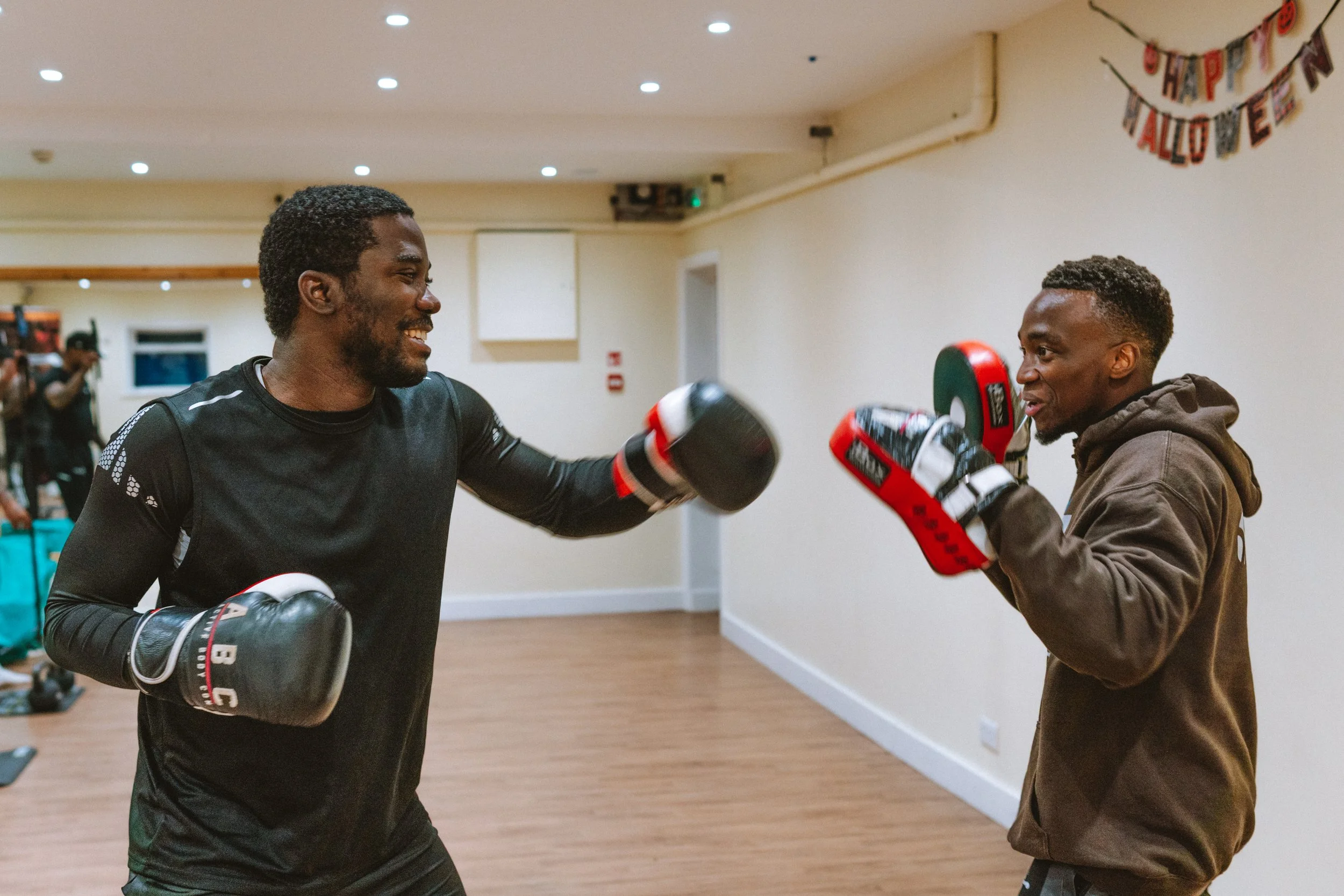 Two men practicing boxing in a gym, one with black gloves and the other with red and black boxing mitts, smiling and fighting.
