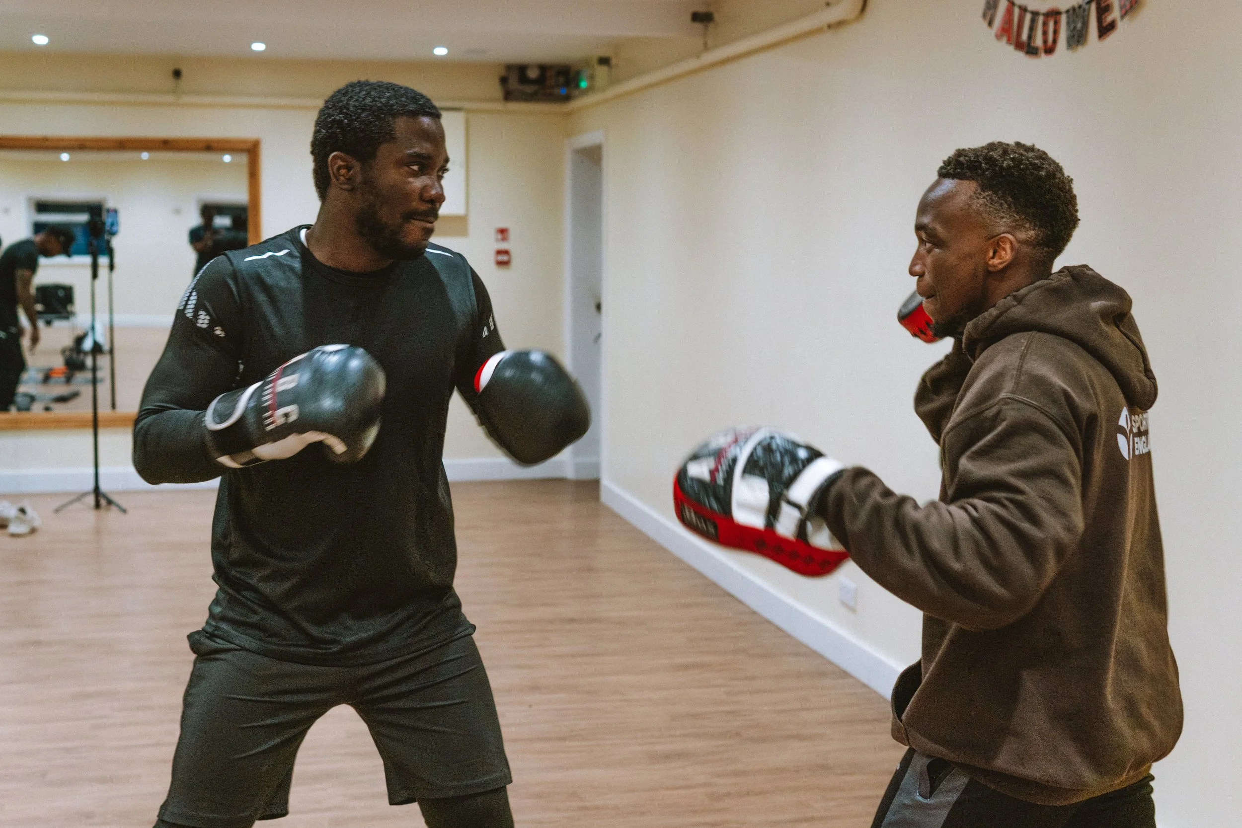Two men in a boxing gym sparring, one in black boxing gloves and athletic wear, the other in a brown hoodie and holding focus mitts.