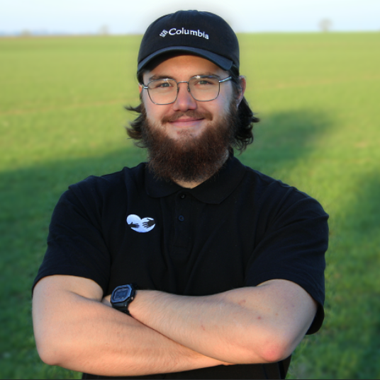 Un homme avec une barbe portant une casquette Columbia et des lunettes, souriant, en plein air dans un champ vert.