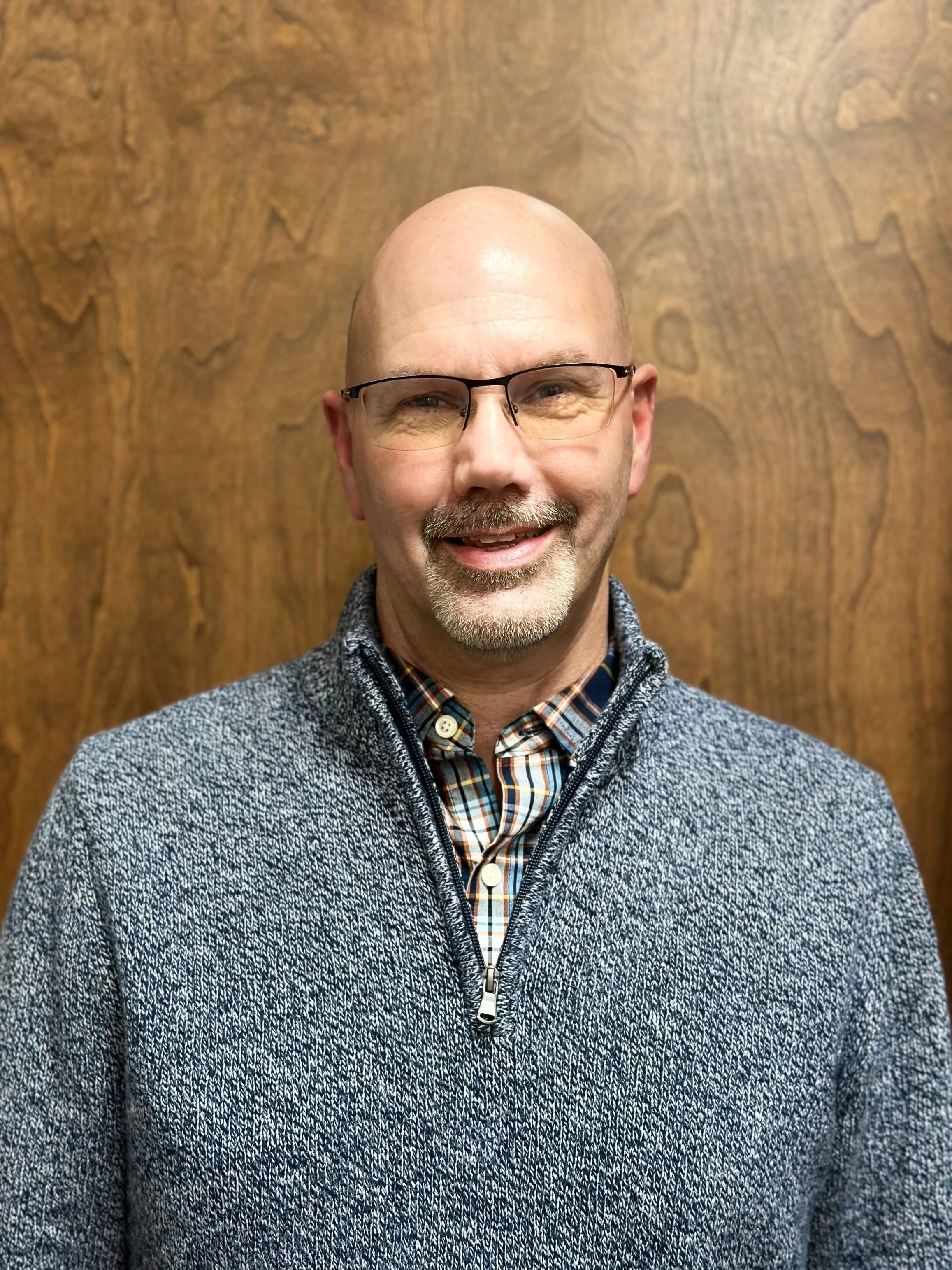 A smiling middle-aged man with glasses, a beard, and a mustache, wearing a plaid shirt and a gray zip-up sweater, standing in front of a wooden background.
