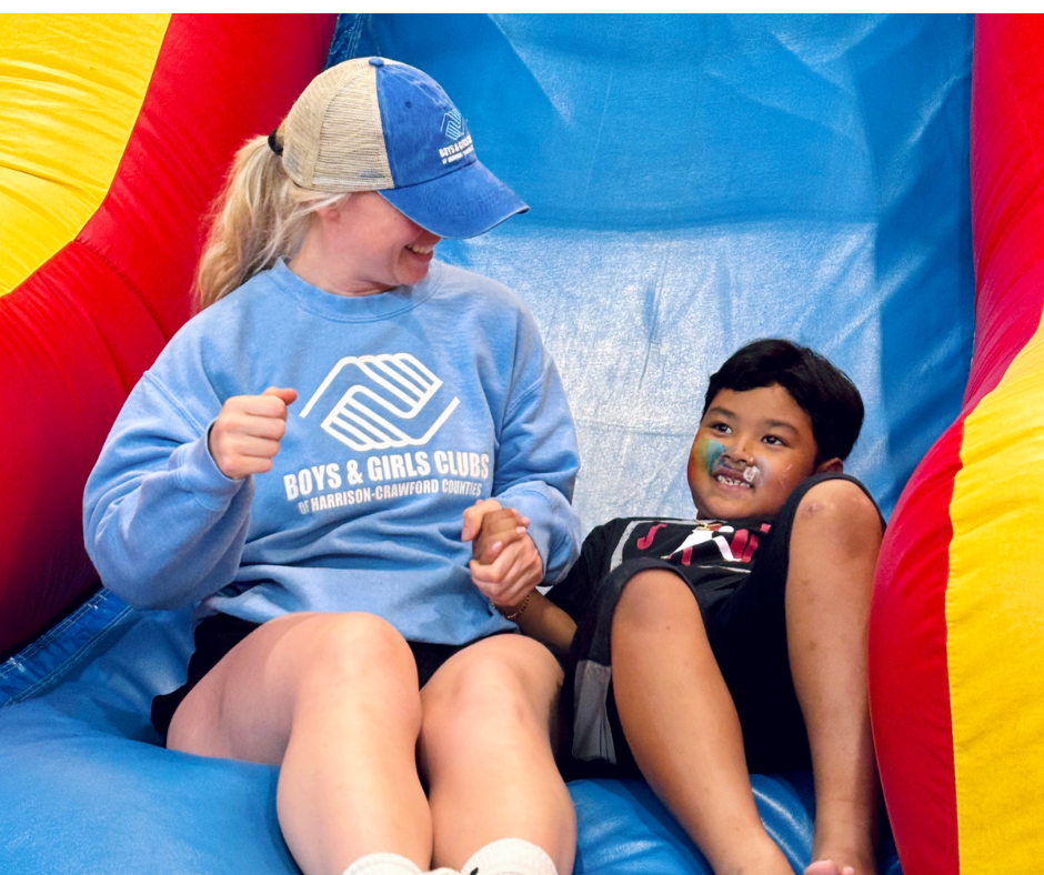An adult woman in Boys & Girls Club attire is going down an inflatable slide with a smiling child with face paint
