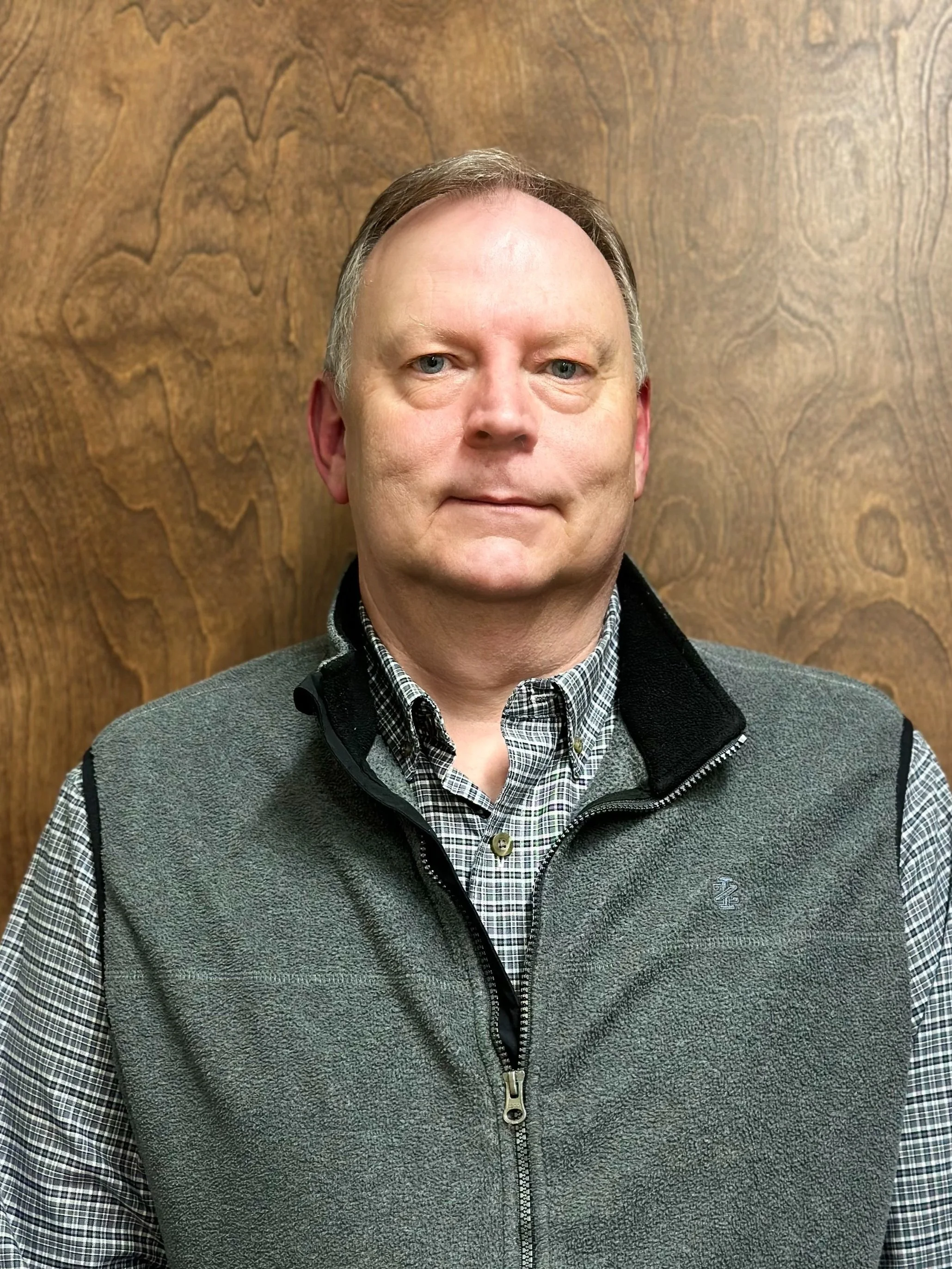 Portrait of a middle-aged man with light skin and short hair, wearing a checkered shirt and a gray vest, standing in front of a wooden background.