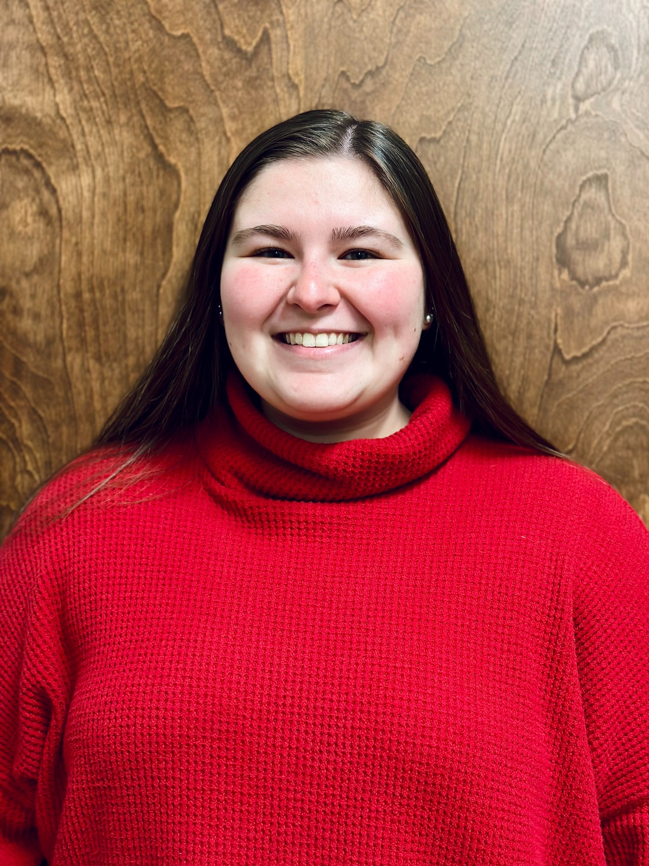 A young woman with long dark hair, smiling, wearing a red turtleneck sweater, standing in front of a wooden wall.