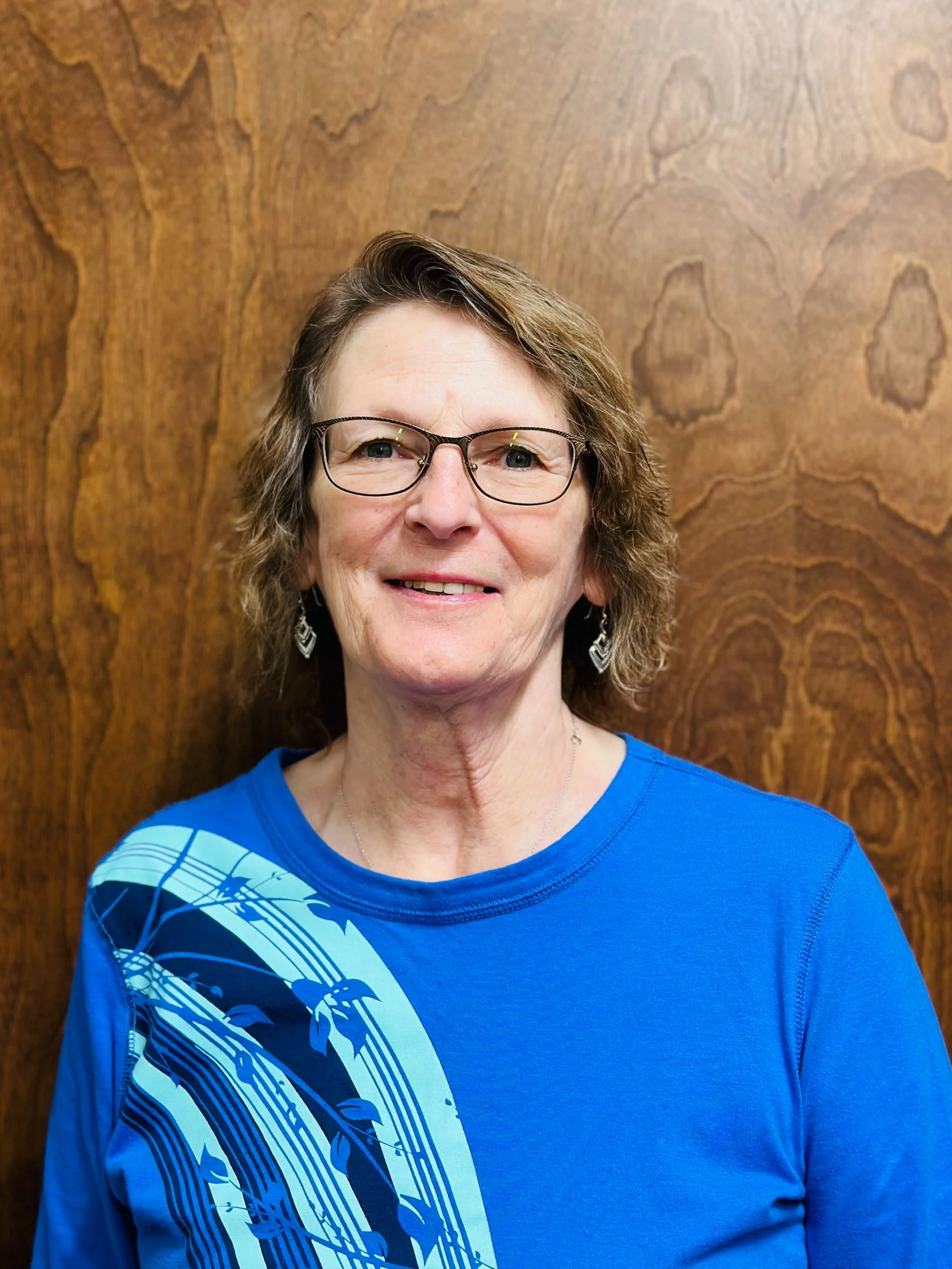 A middle-aged woman with glasses and earrings smiling in front of a wood-paneled wall.