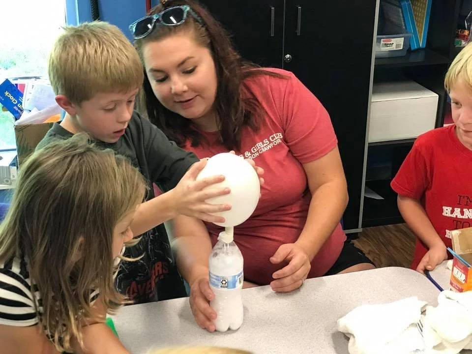 An adult woman in a Boys & Girls Club shirt assists a young boy and girl with a science experiment