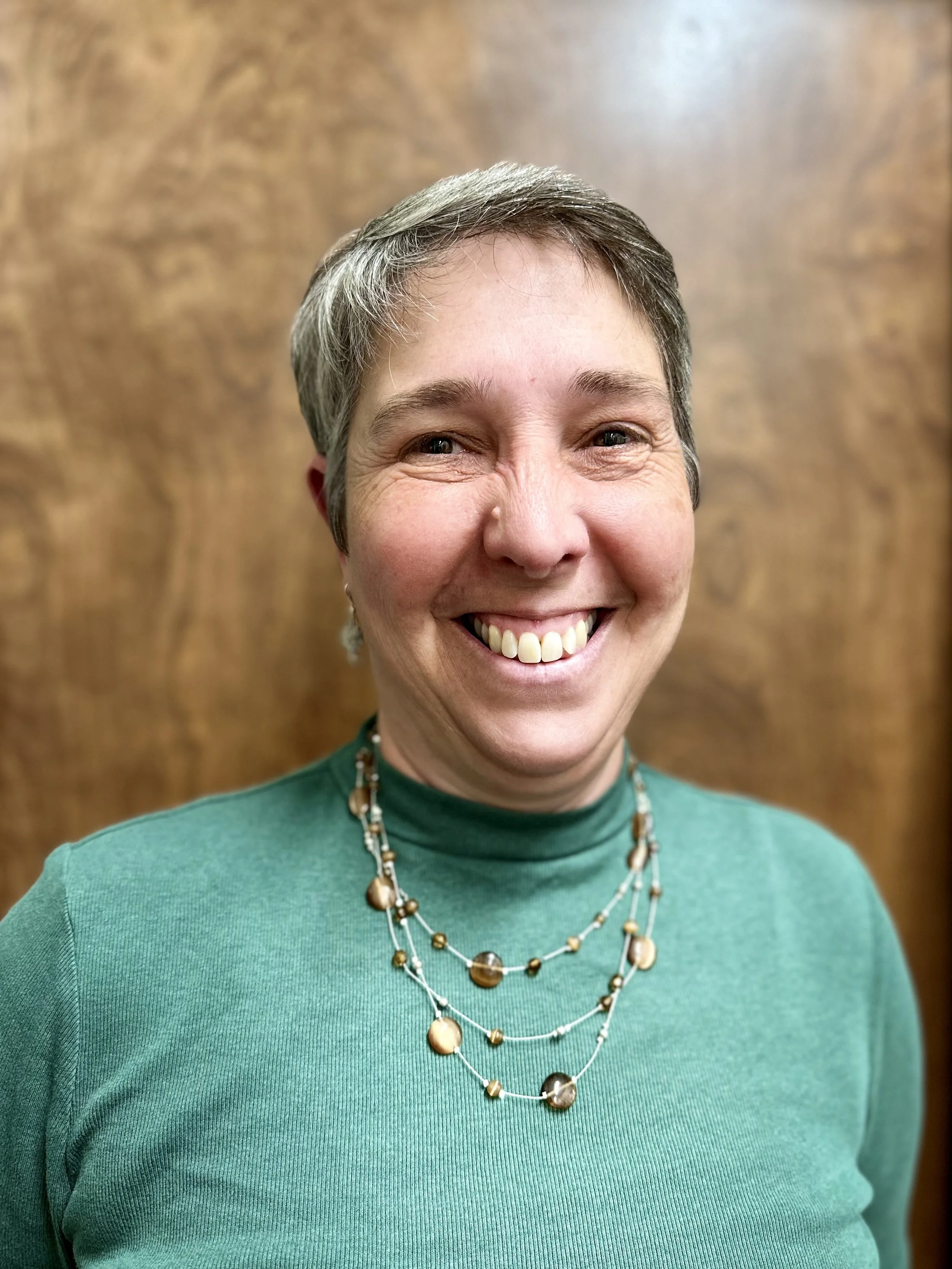 A smiling woman with short gray hair, wearing a green turtleneck and layered pearl necklaces, standing in front of a wooden background.