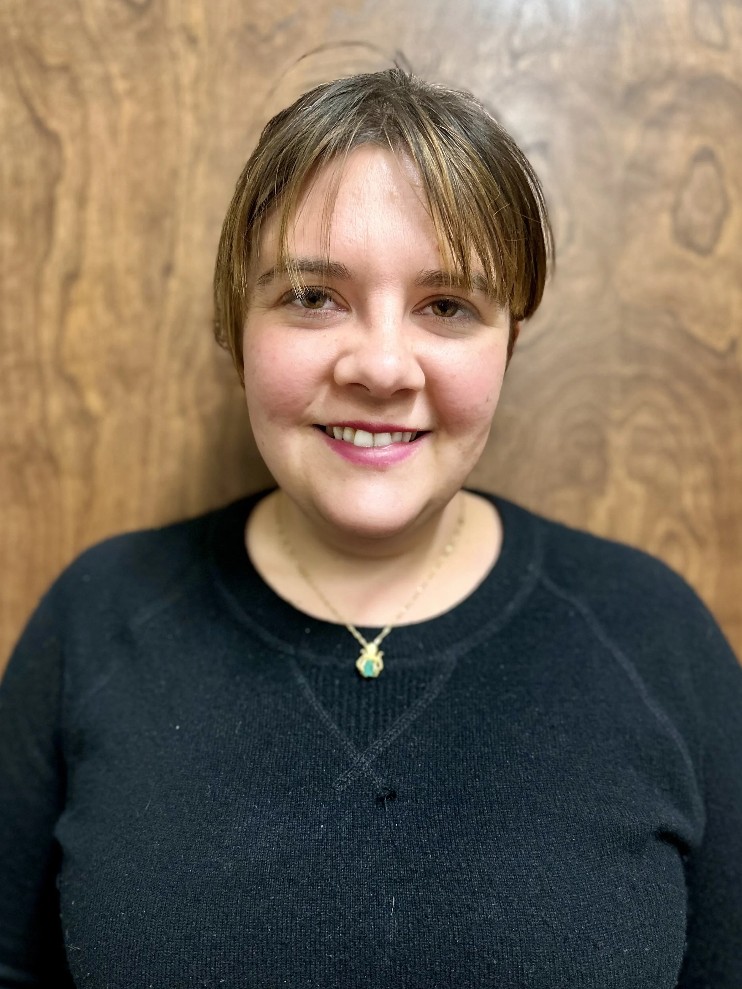 A woman with short brown hair smiling, wearing a black top and a gold necklace with a small pendant, standing in front of a wooden background.