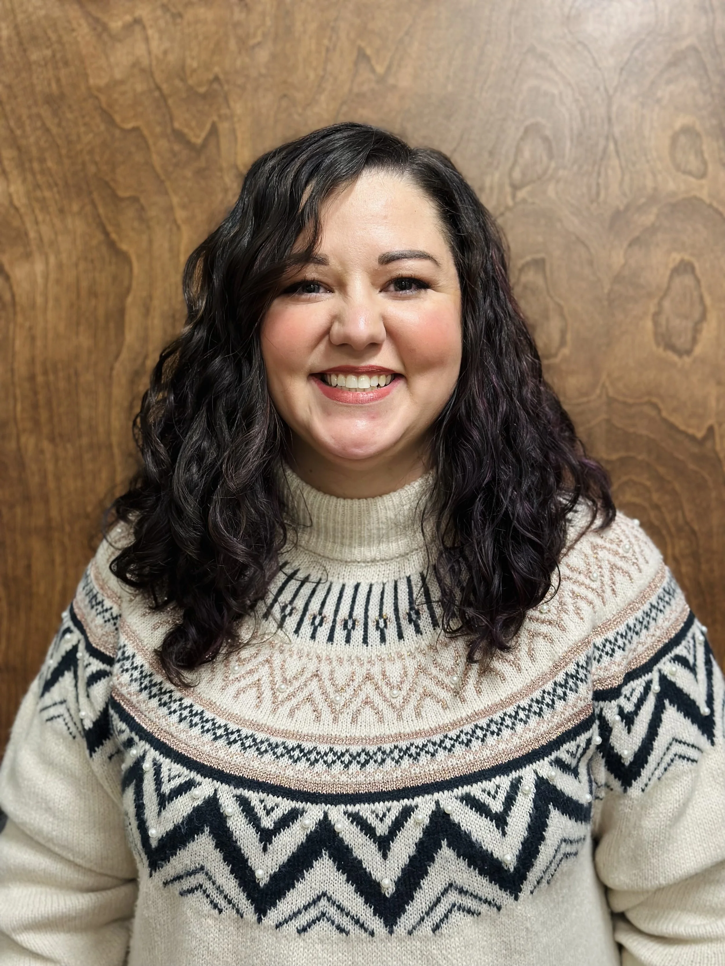 Portrait of a smiling woman with dark, curly hair, wearing a cream-colored sweater with a black and pink geometric pattern, standing in front of a wooden background.