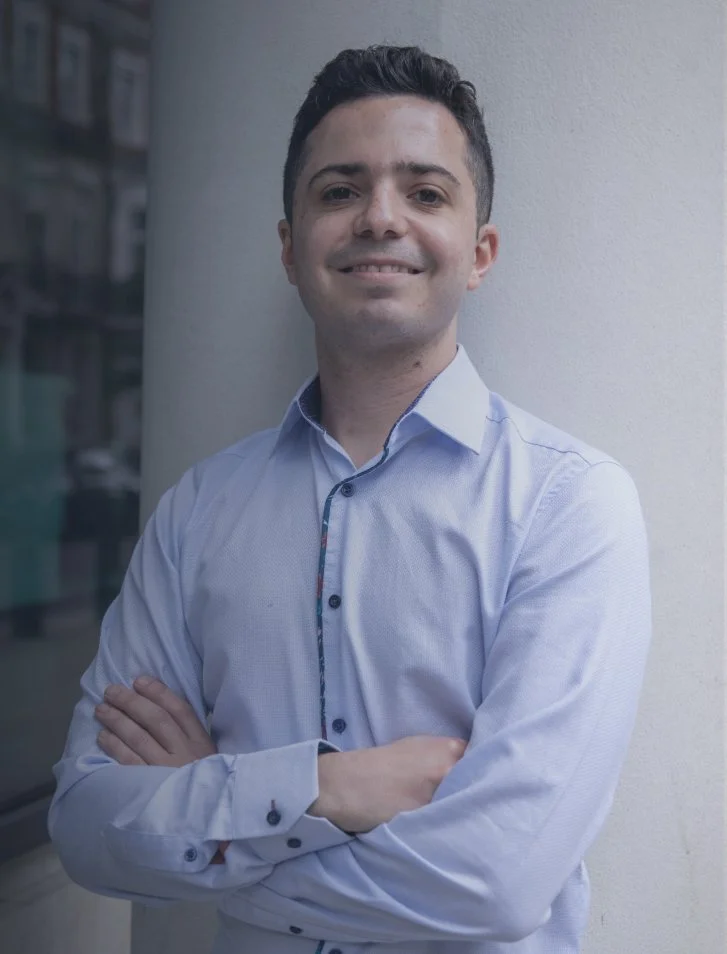 A young man with short dark hair, smiling, wearing a light blue dress shirt with dark buttons, standing outdoors next to a white column, with an urban street reflected in a window behind him.